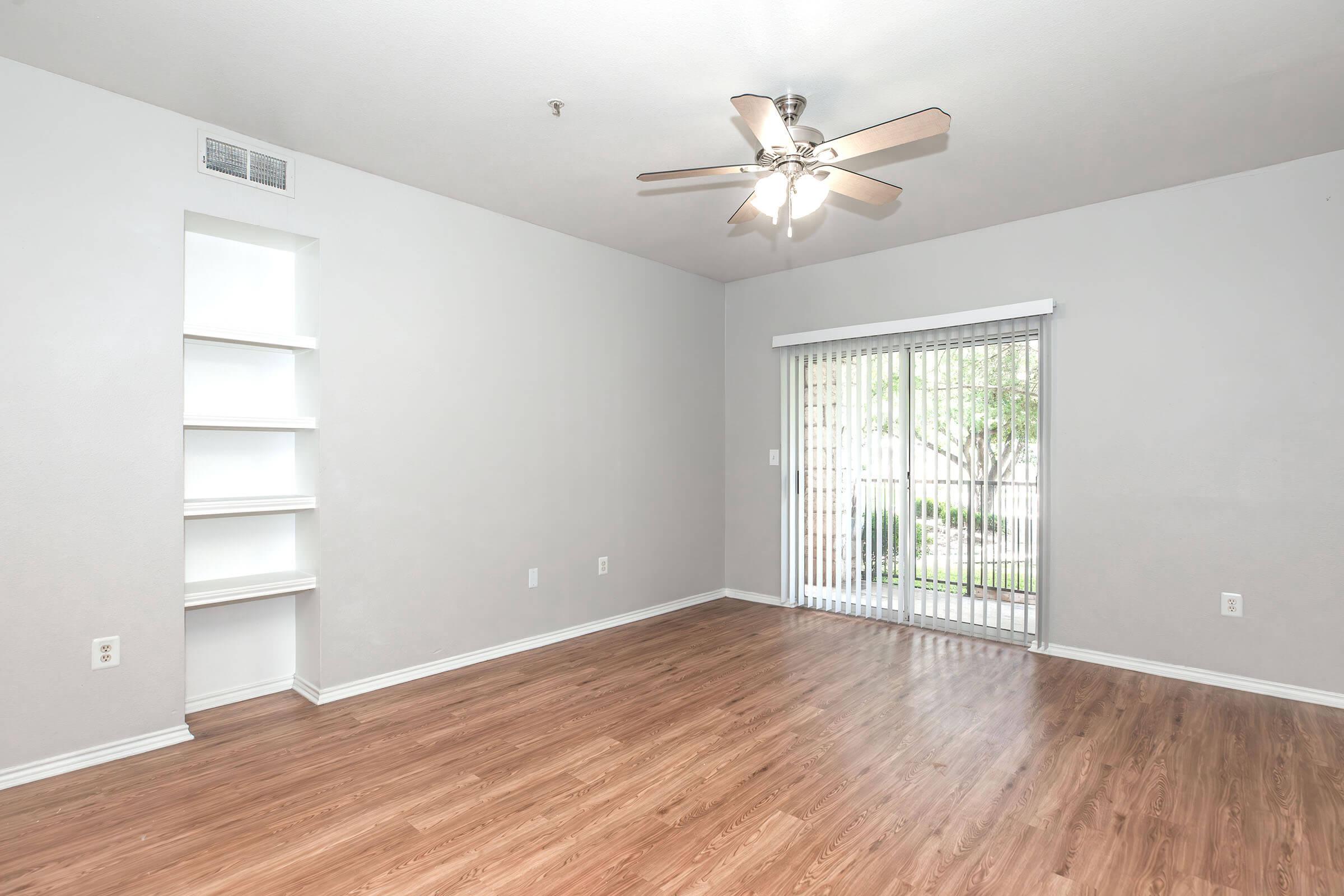 An empty living room featuring light-colored walls, a ceiling fan with light, and a sliding glass door that opens to a view of greenery outside. There is a small built-in shelf on one wall, and the floor is made of light wood laminate. The space is bright and airy.