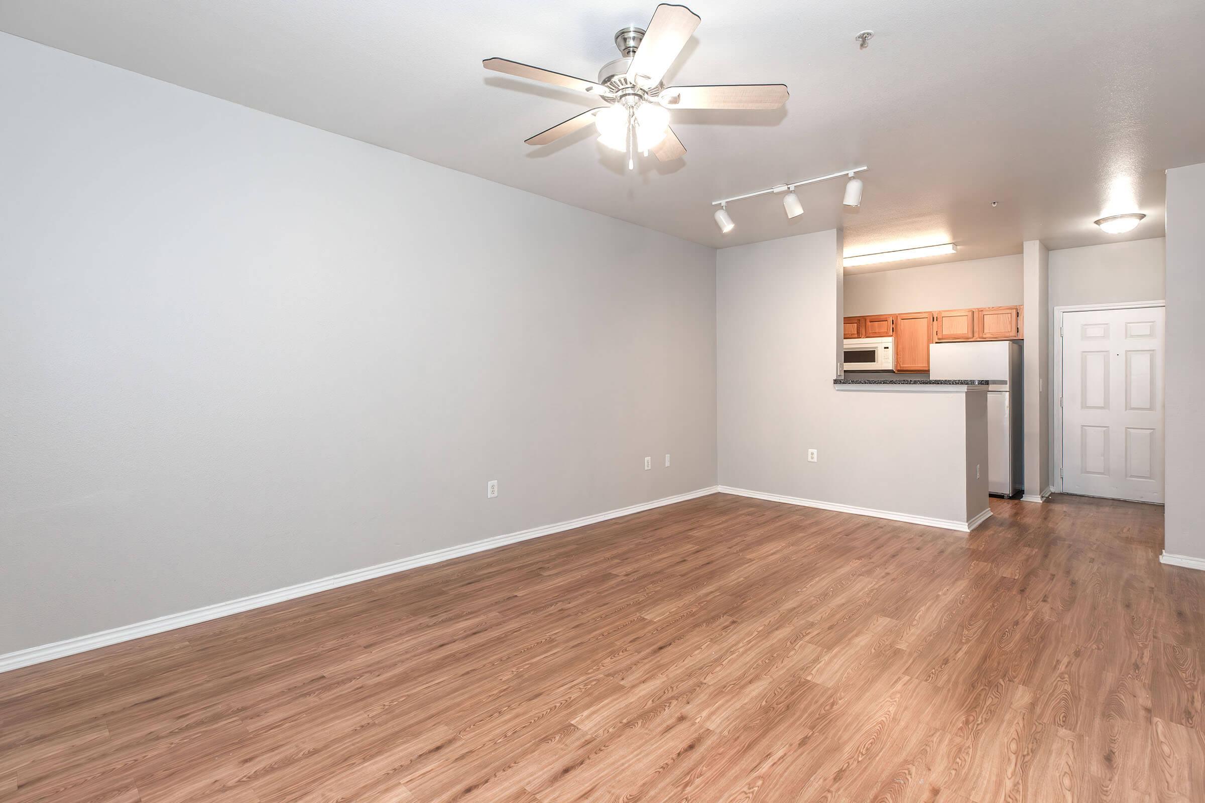 A spacious, modern living area featuring light wood-like flooring, a ceiling fan with lights, and pale gray walls. A kitchen area with wooden cabinets and appliances is visible in the background, while a doorway leads to another room. The space is well-lit and ready for furniture arrangement.