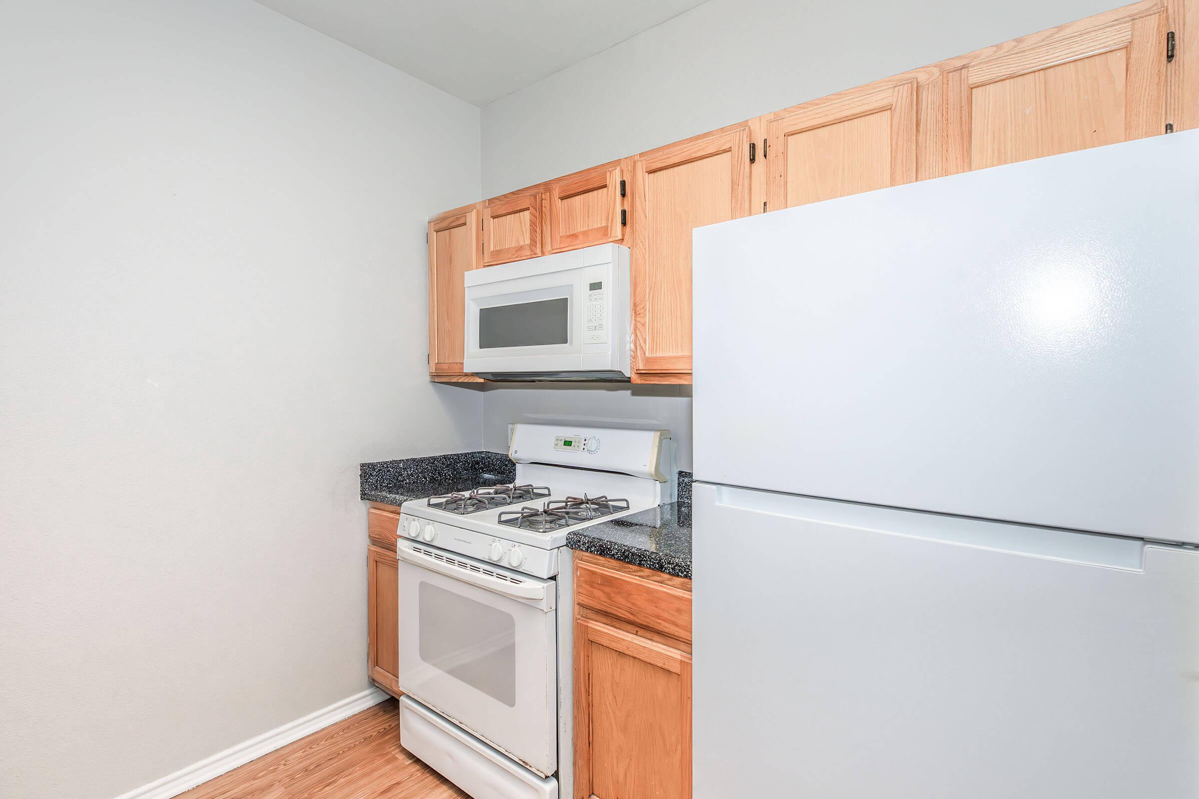 A kitchen featuring light wood cabinets, a white microwave above a white stove, and a refrigerator. The countertop is dark granite, and the flooring is a light wood finish. The walls are painted a neutral color, creating a modern and spacious appearance.