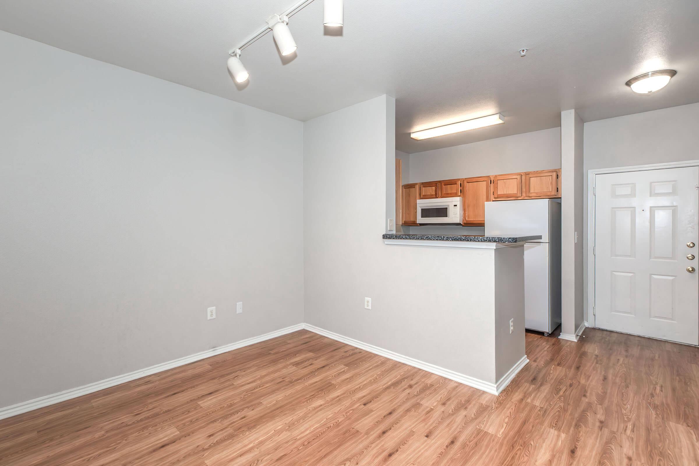 A spacious, empty apartment with light-colored walls, a wooden floor, and a small kitchen area visible in the background. There are wooden cabinets, a refrigerator, and a door leading outside. Bright overhead lights illuminate the room, creating a clean and inviting atmosphere.