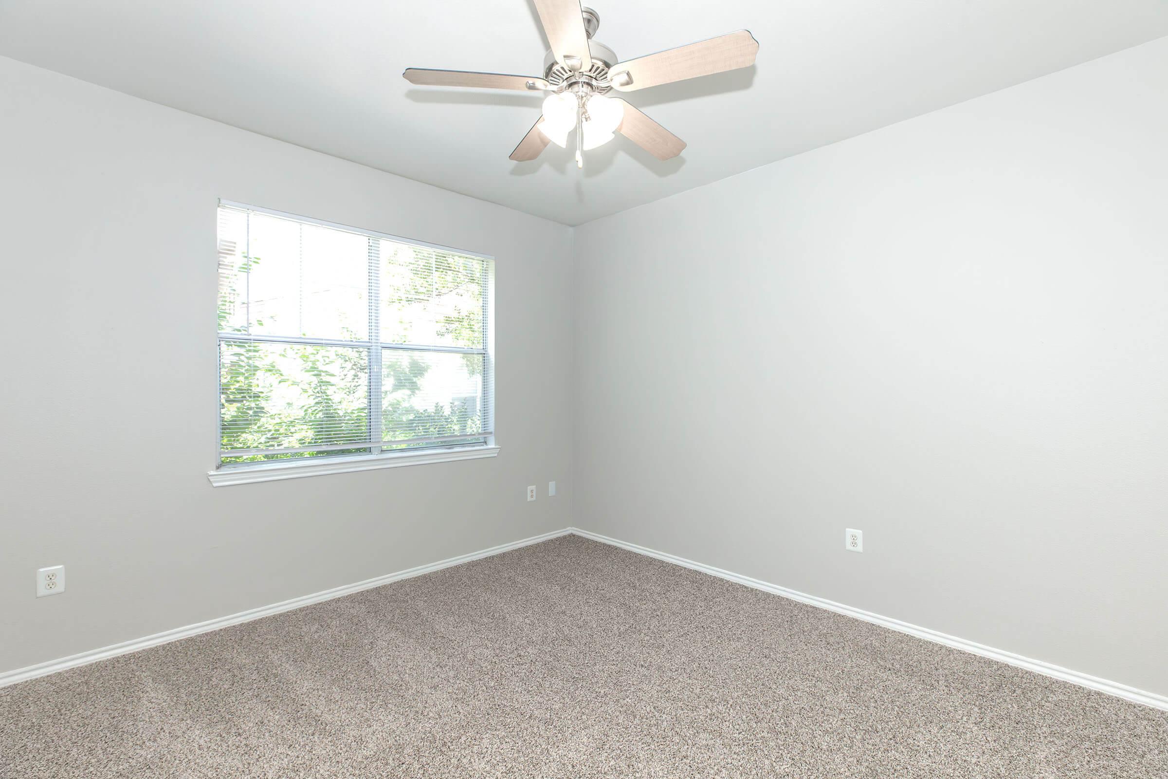 A vacant, well-lit room featuring light gray walls, a ceiling fan with wooden blades, and carpeted flooring. There is a large window allowing natural light, overlooking greenery outside. The space appears clean and ready for furnishing.