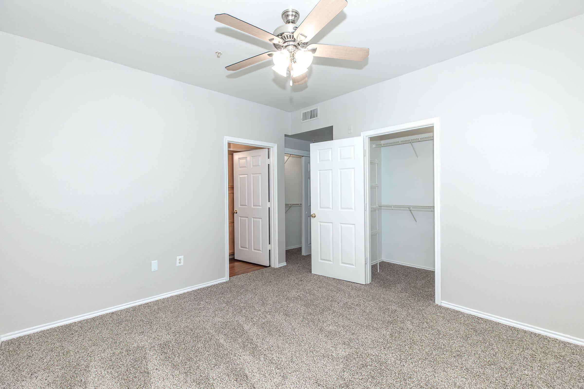 A spacious room featuring light gray walls and a ceiling fan with wooden blades. Two doors lead to adjacent areas, while a closet with shelves is visible on the right. The floor is covered in soft, beige carpet, creating a cozy and inviting atmosphere.