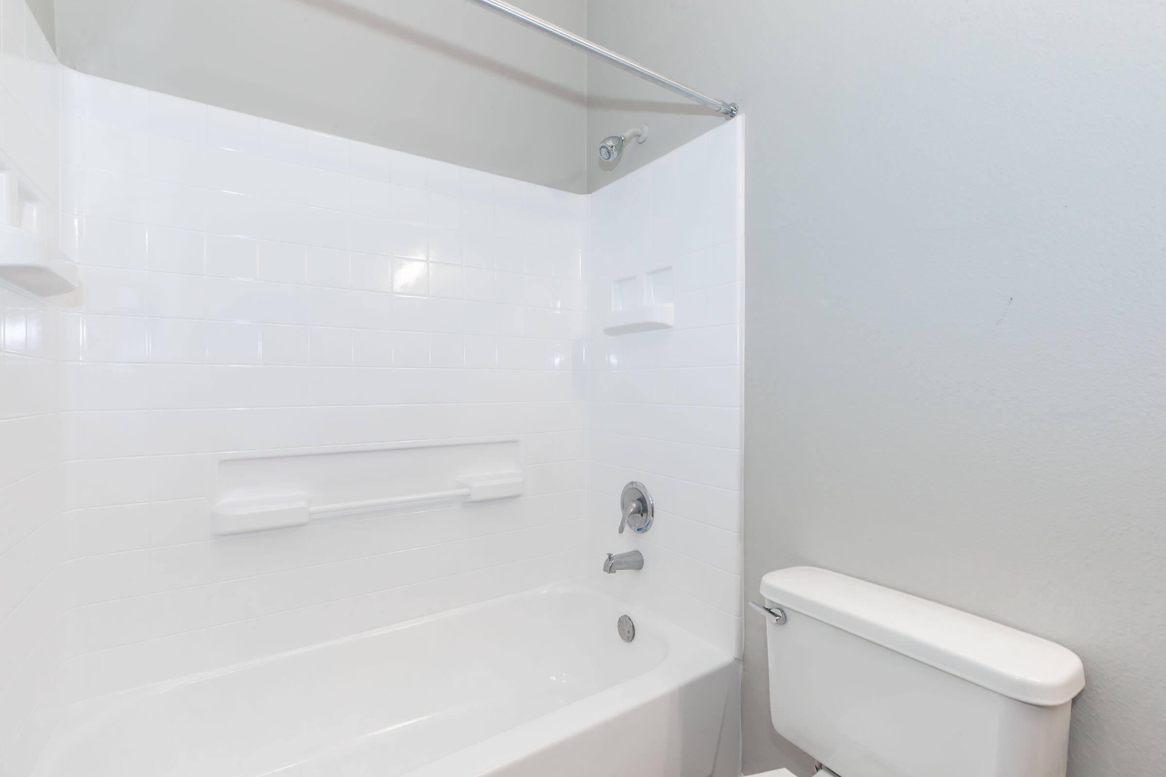 A clean, modern bathroom featuring a white bathtub with a shower head, a glass shower curtain rod, and a wall-mounted grab bar. The walls are painted light gray, and there is a white toilet beside the bathtub. The overall design is minimalistic and bright.