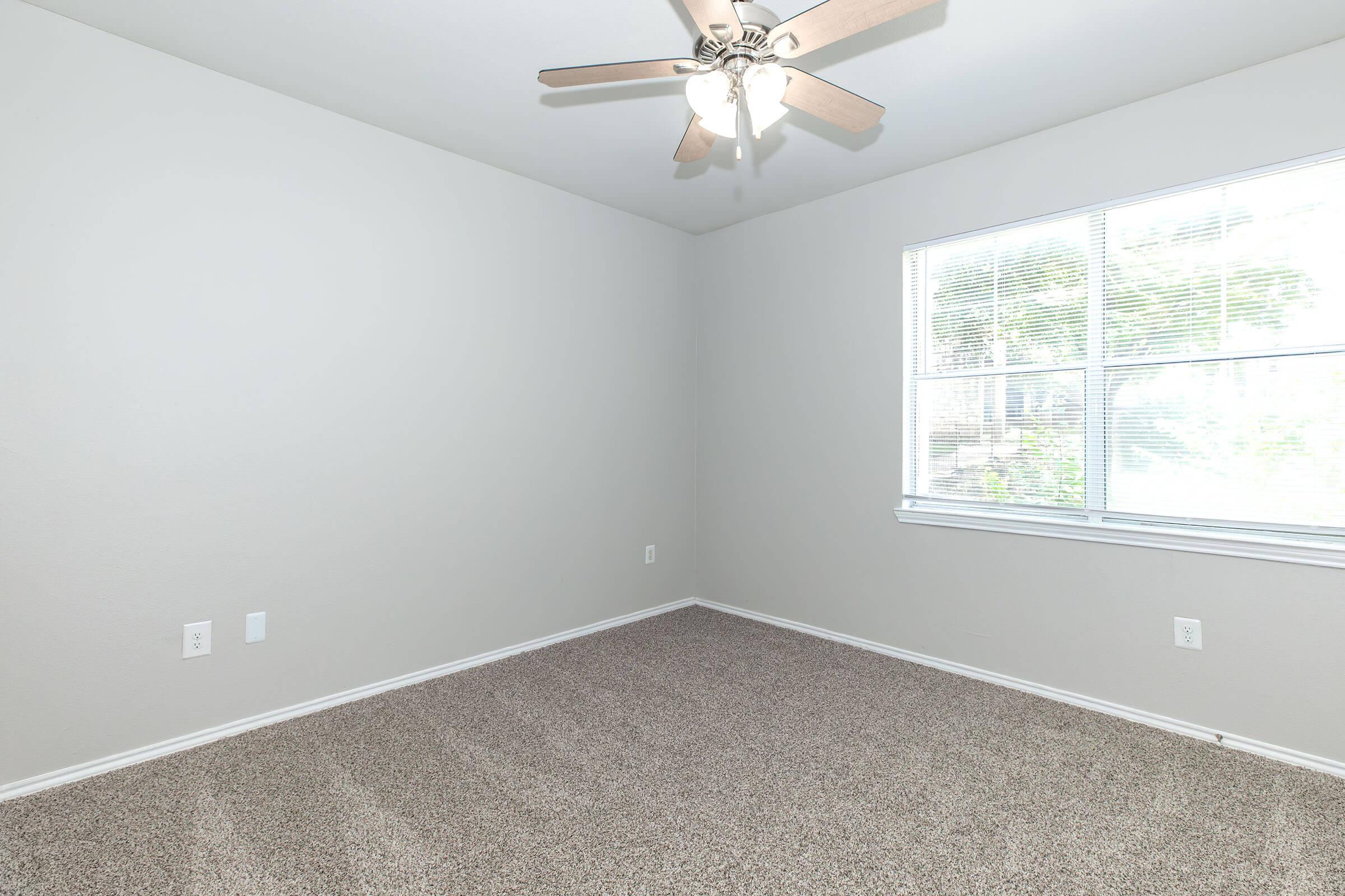 Empty room with beige carpeting and light gray walls. A ceiling fan is mounted in the center, and there is a large window with blinds that allows natural light to enter. The room is unfurnished, creating a spacious, minimalist atmosphere.