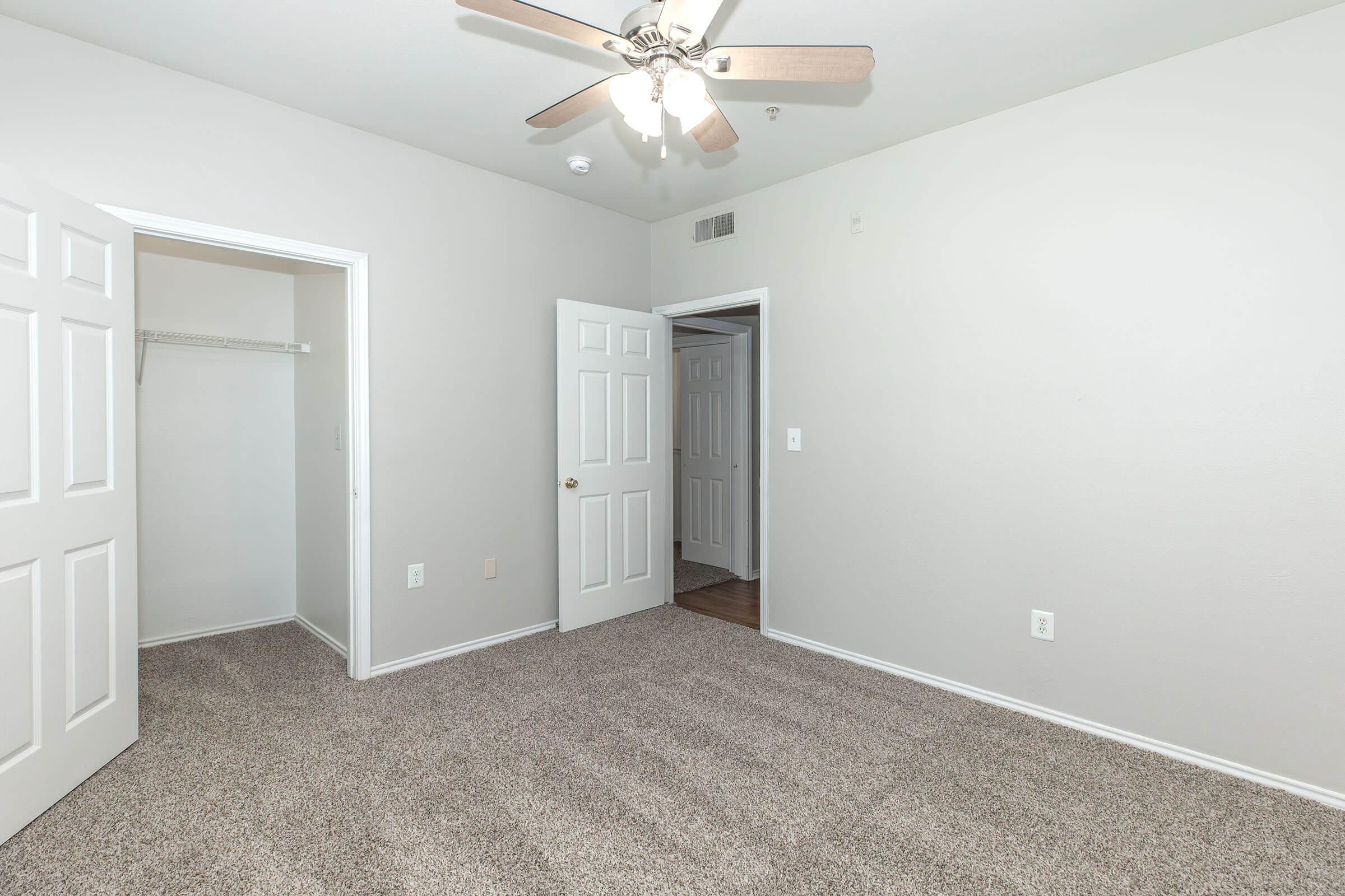 A clean, empty room with light-colored walls and carpeted flooring. There is a ceiling fan, two closed doors leading to separate spaces, and a closet area visible on the left. The room is well-lit, providing a spacious and airy atmosphere.