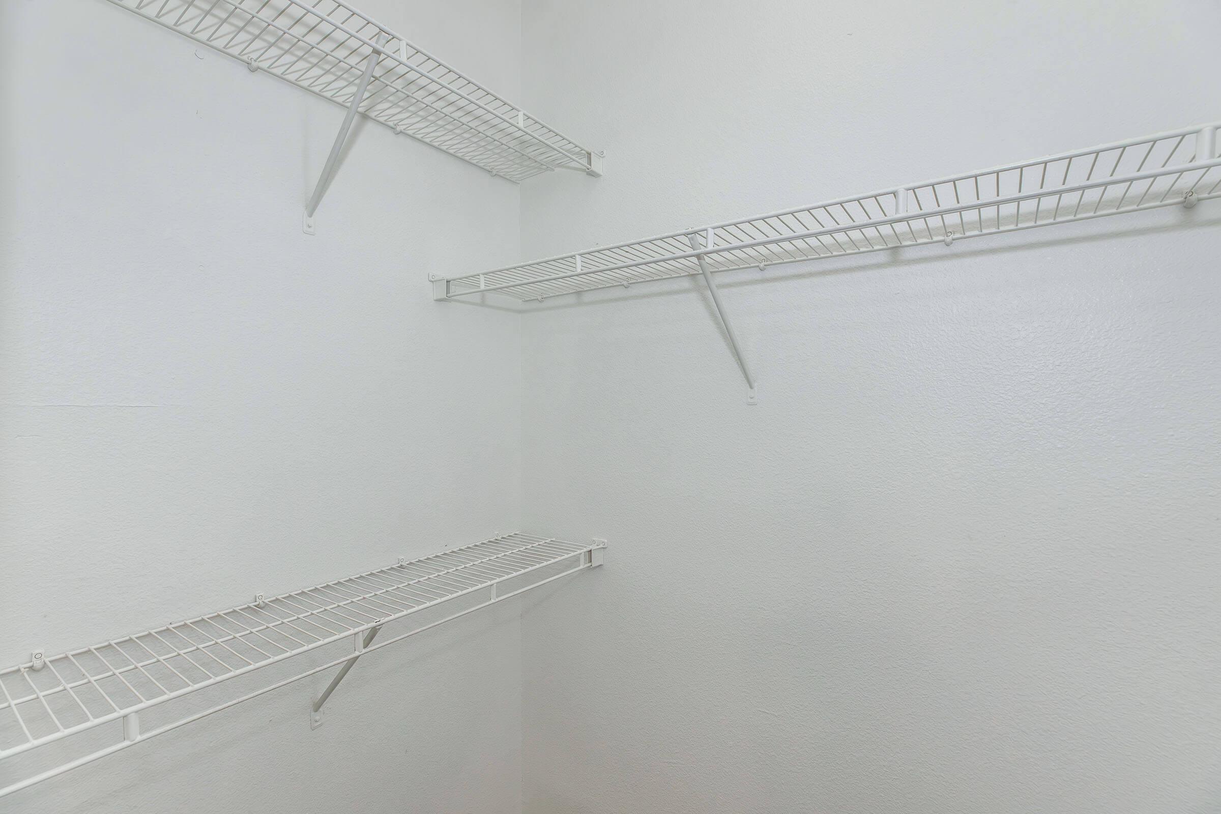 Empty white storage closet featuring several metal wire shelving units mounted on the walls. The interior is well-lit, showcasing the clean, minimalist space with no items on the shelves.