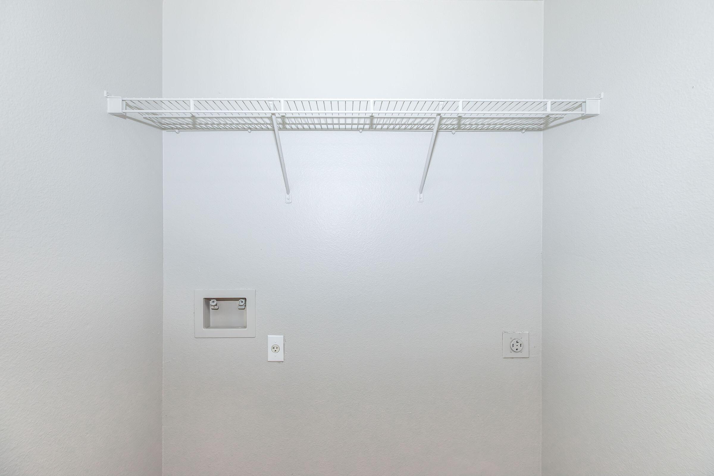 A blank wall with a wire shelving unit mounted near the top. The wall is painted white, and there is a small section visible with a towel rack and electrical outlets below the shelf. Overall, the space appears clean and minimalistic.