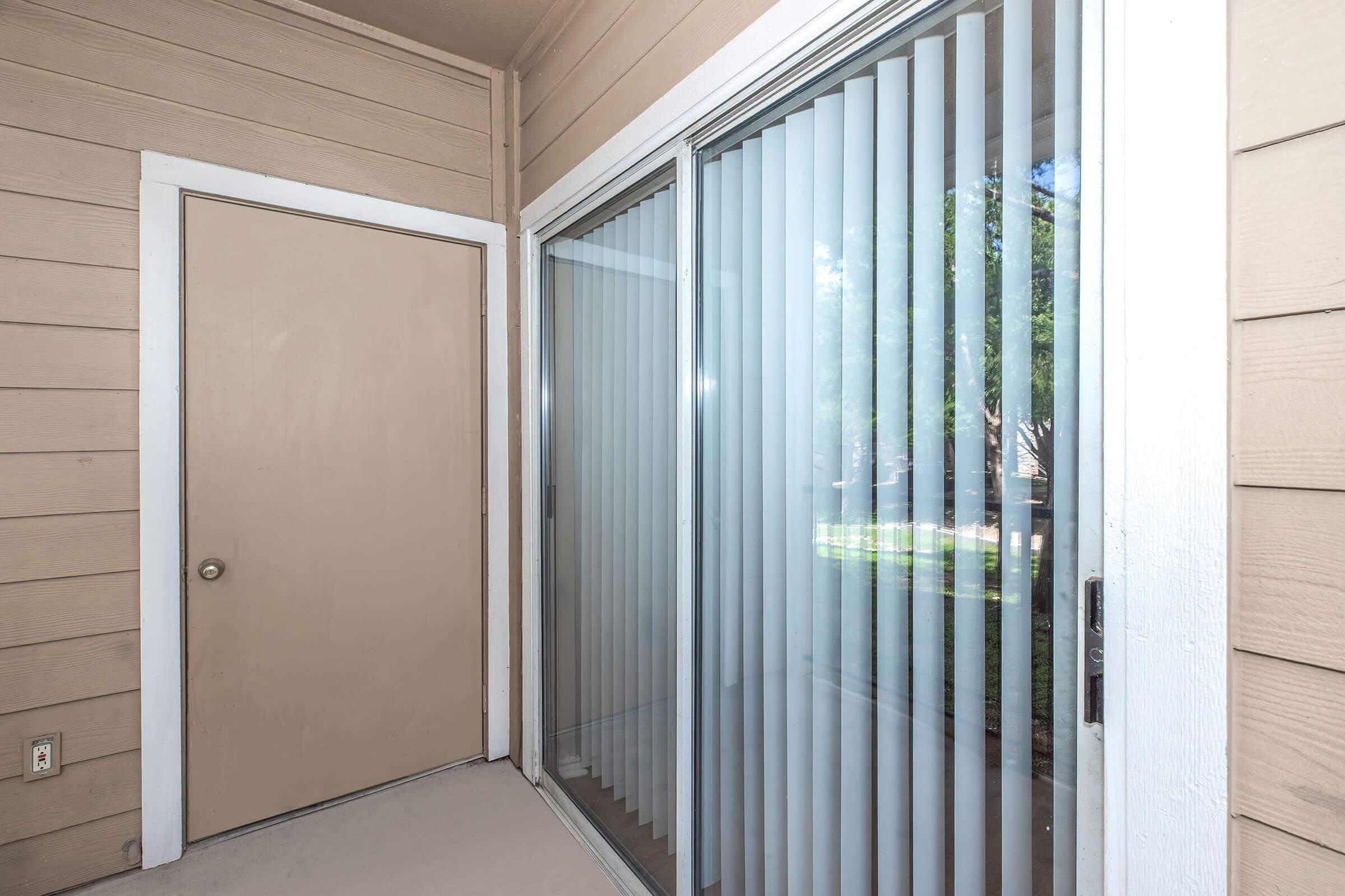 A corner view of a patio or balcony area featuring a closed door and large sliding glass door with vertical blinds. The wall is light brown, and there is greenery visible outside through the glass. The floor is concrete, and the setting appears well-lit.