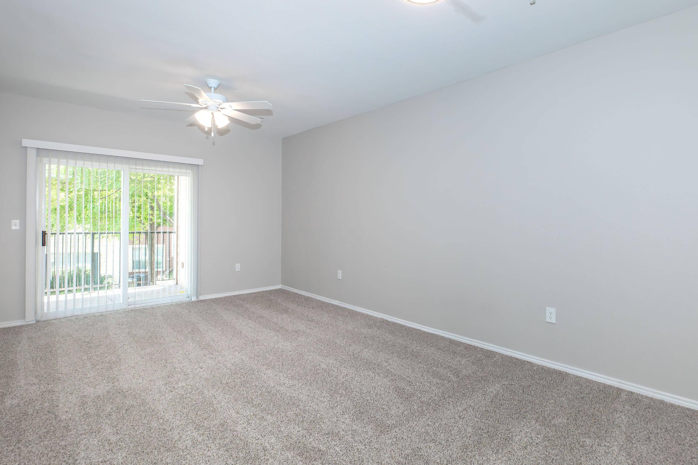 Empty room with light gray walls and beige carpet, featuring a ceiling fan. There is a sliding glass door leading to a balcony, with natural light coming in. The space is unfurnished, creating a clean and open atmosphere.