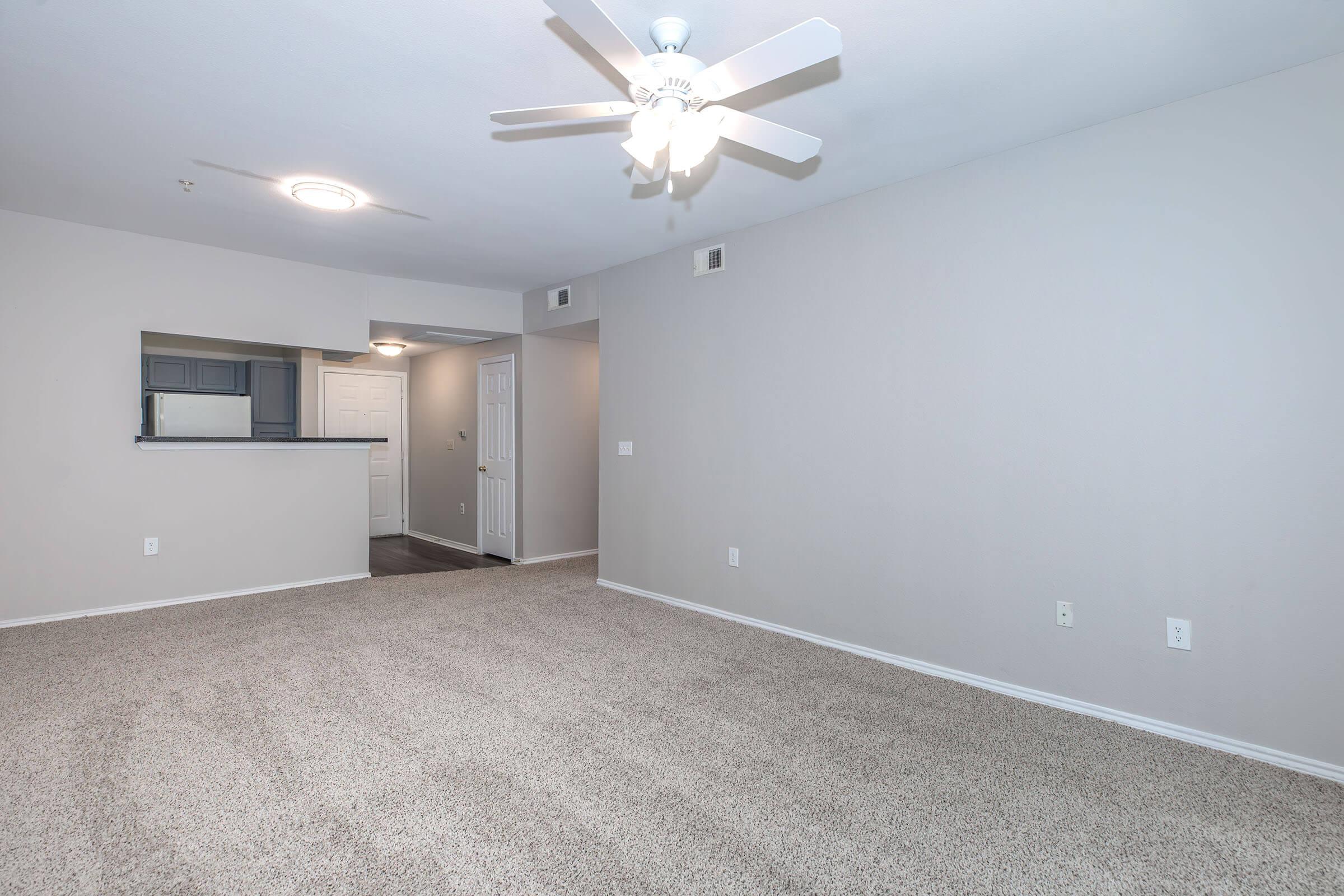 An empty apartment living room featuring light gray walls, a ceiling fan, and beige carpet. To the left is a partial view of a kitchen area with a bar counter, and a doorway leading to another room is visible. There are no furnishings or decorations in the space, giving it a spacious and clean appearance.