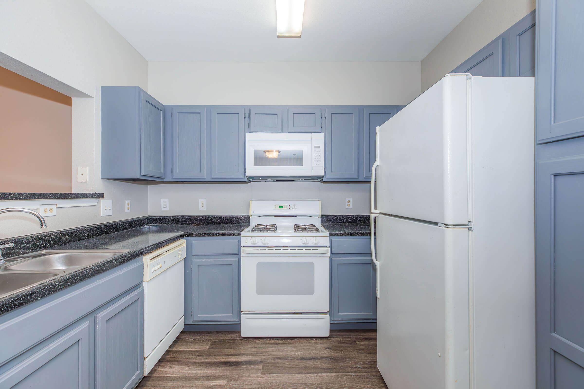 A modern kitchen with blue cabinet doors, a white refrigerator, a gas stove, and a built-in microwave. The countertops are dark and feature a double sink, with light-colored walls and laminate flooring. The space is well-lit with overhead lighting.