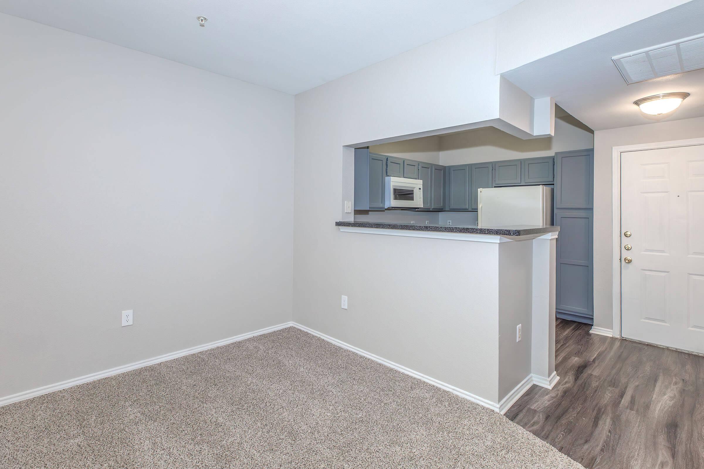 Interior view of a modern apartment showing a small living area with beige carpet, light gray walls, and a kitchen with blue cabinetry visible through an open counter. A white door leads to the entrance, and a white refrigerator is seen in the kitchenette area. Bright lighting enhances the open space.
