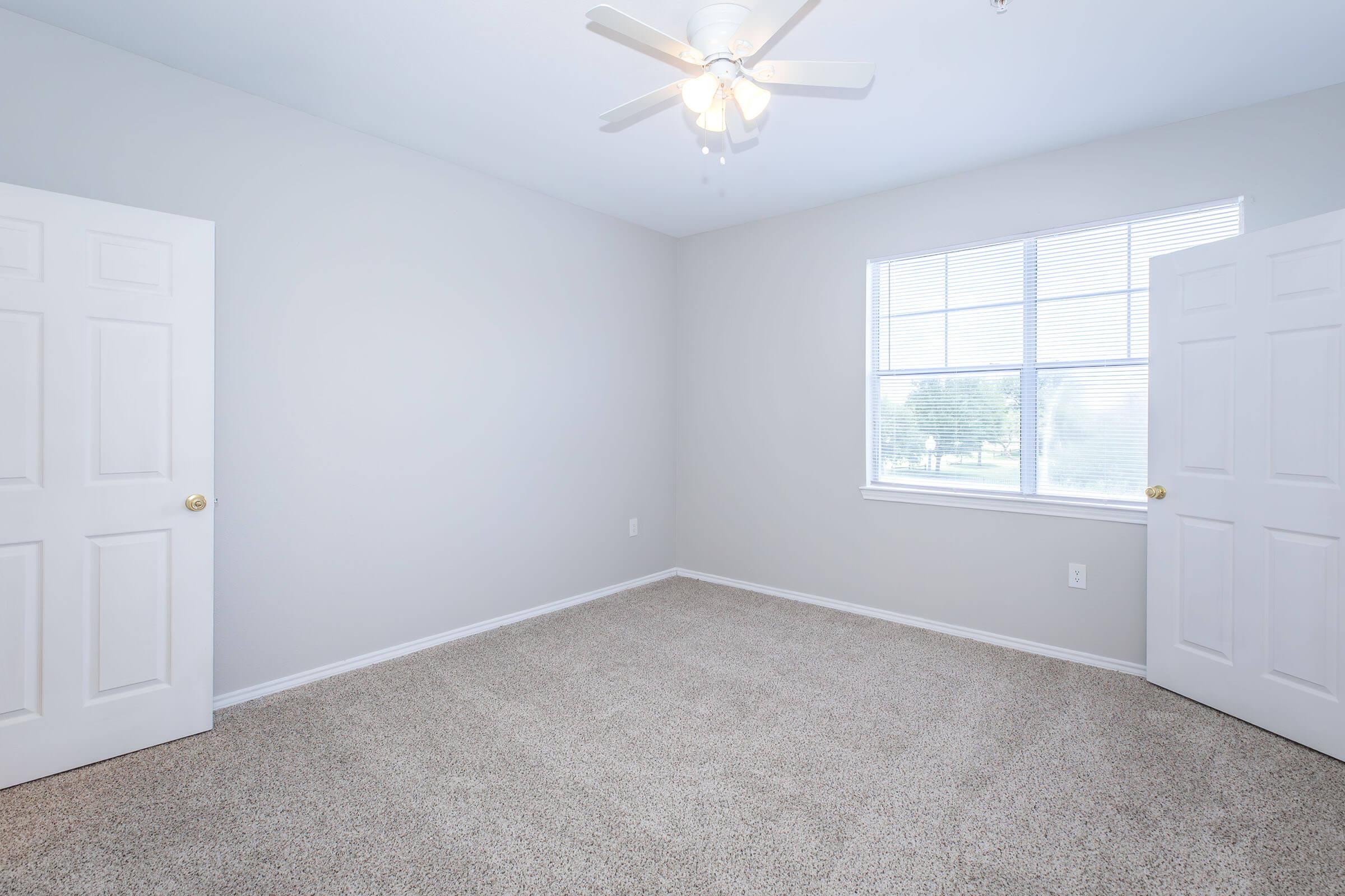 A clean, empty bedroom featuring beige carpet and light gray walls. There is a ceiling fan with white blades, and two open doors leading to the hallway. A window with blinds allows natural light to fill the space, creating an airy atmosphere.