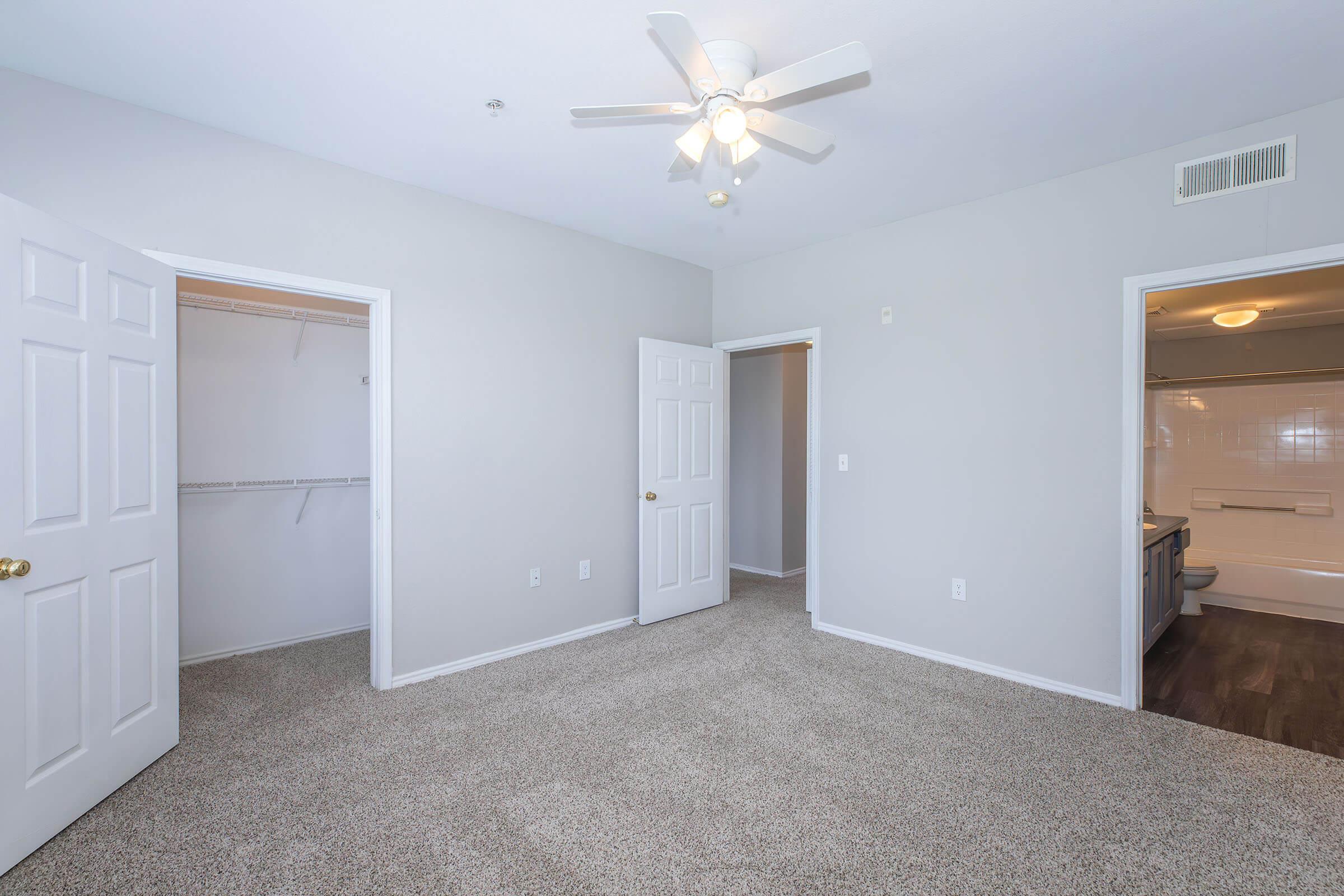A well-lit, spacious room with light gray walls and a ceiling fan. Two open double doors lead to a closet and a bathroom. The floor is covered in soft beige carpet, creating a cozy atmosphere. The doorway to the bathroom is visible in the background, showcasing a clean and simple design.