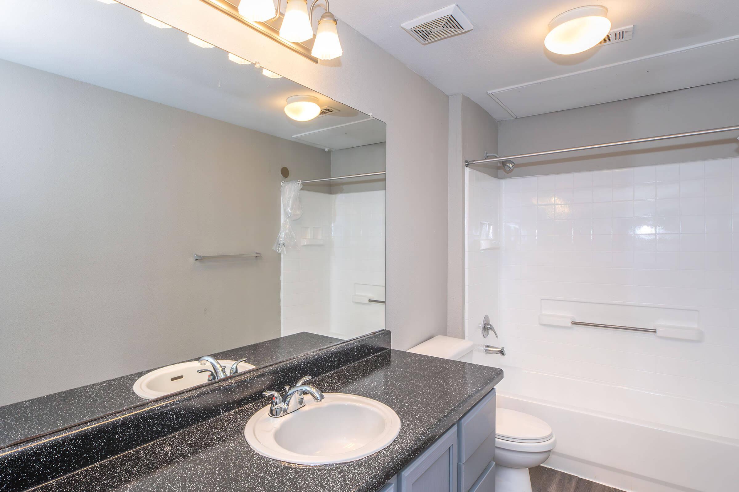 A clean, modern bathroom featuring a dark granite countertop with a double sink, a large mirror, and a bathtub with a white tile surround. The walls are painted a light gray, and there is a towel rack and overhead lighting. The floor is tiled, creating a contemporary look.