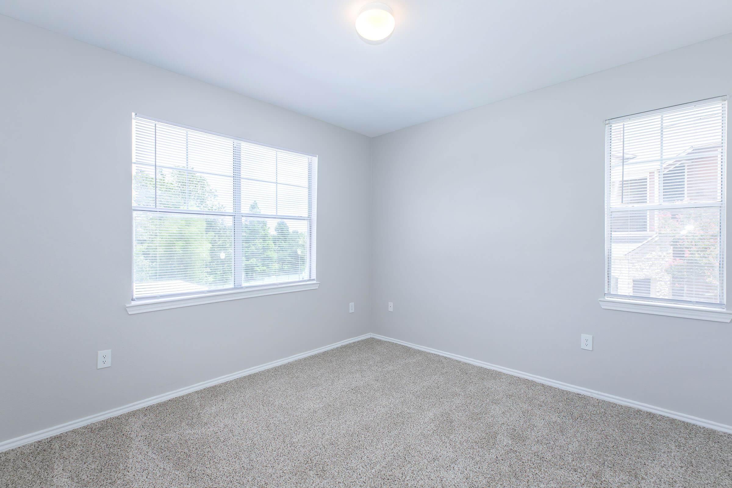 Empty room with light gray walls, two windows allowing natural light, and beige carpet flooring. The space appears clean and spacious, with no furniture or decorations present. The windows provide a view of the outdoors.