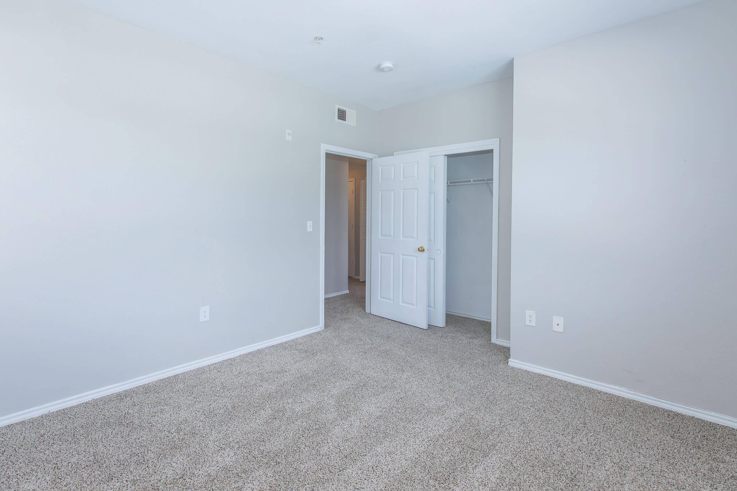 A spacious, empty room with light gray walls and carpet flooring. There are two open doors leading to a closet space and another area. The room features a neutral color palette, allowing for versatile decorating options. Natural light may come through windows not visible in the image.