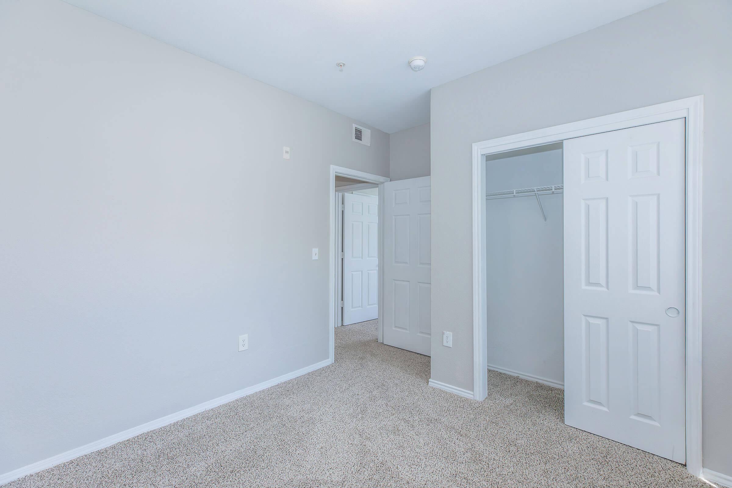 Empty room featuring light gray walls and carpeted flooring. There are two closed white doors; one leads to a closet while the other appears to open into a hall. The space is well-lit, with a neutral and modern aesthetic.