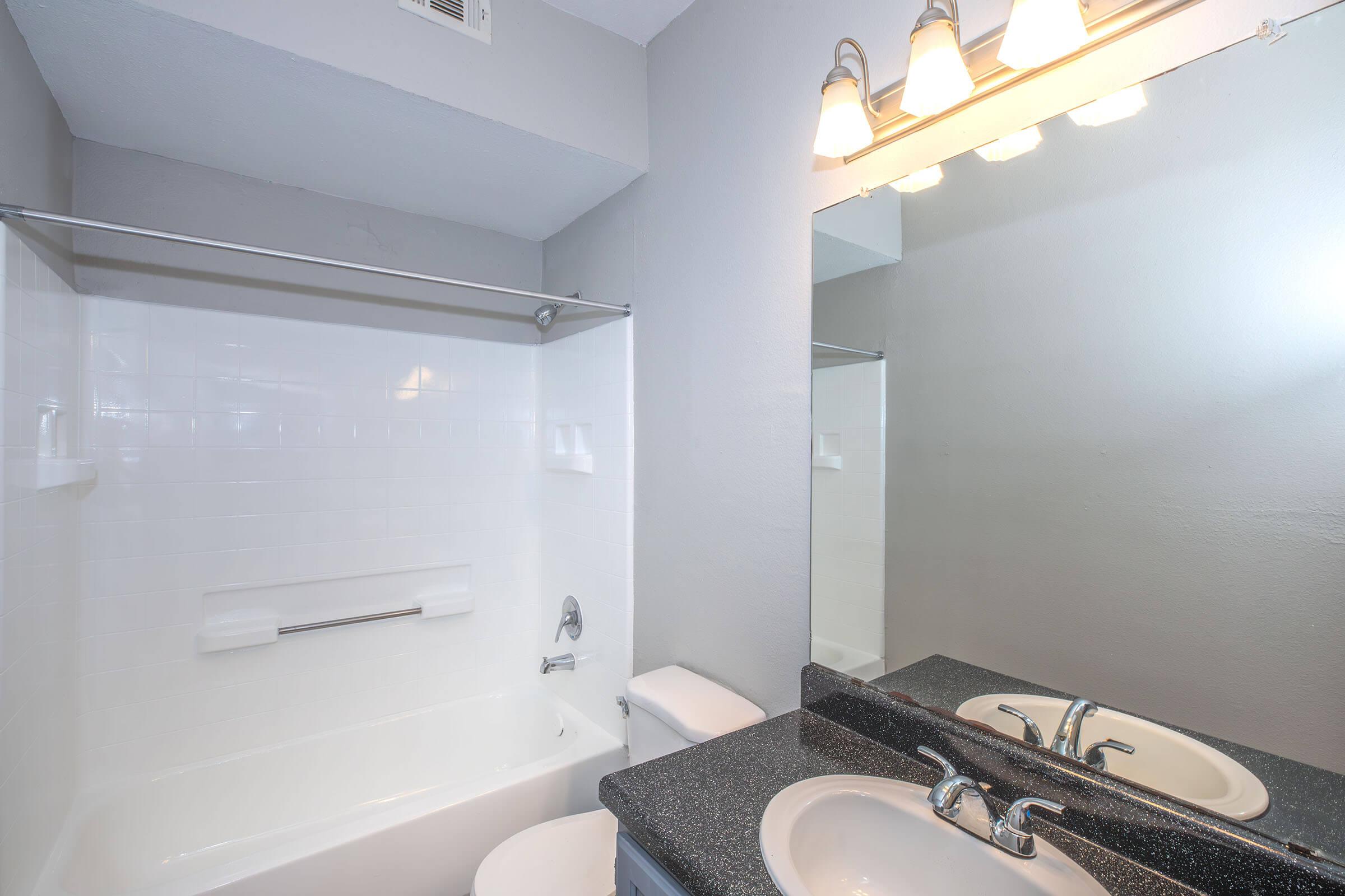 A clean and modern bathroom featuring a white bathtub with a shower, a single sink with a dark countertop, a toilet, and a large mirror above the sink. The walls are painted in a light gray, and there are three light fixtures above the mirror.