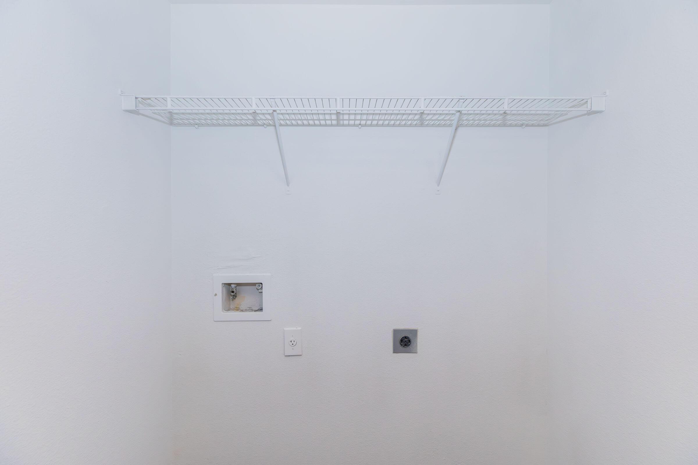 Empty room with white walls, featuring a wire shelf mounted on the wall. There is a small electrical outlet and a box in the wall, indicating potential for future installation of appliances or utilities. The space is uncluttered and minimalistic, suggesting it could be used for storage or laundry.