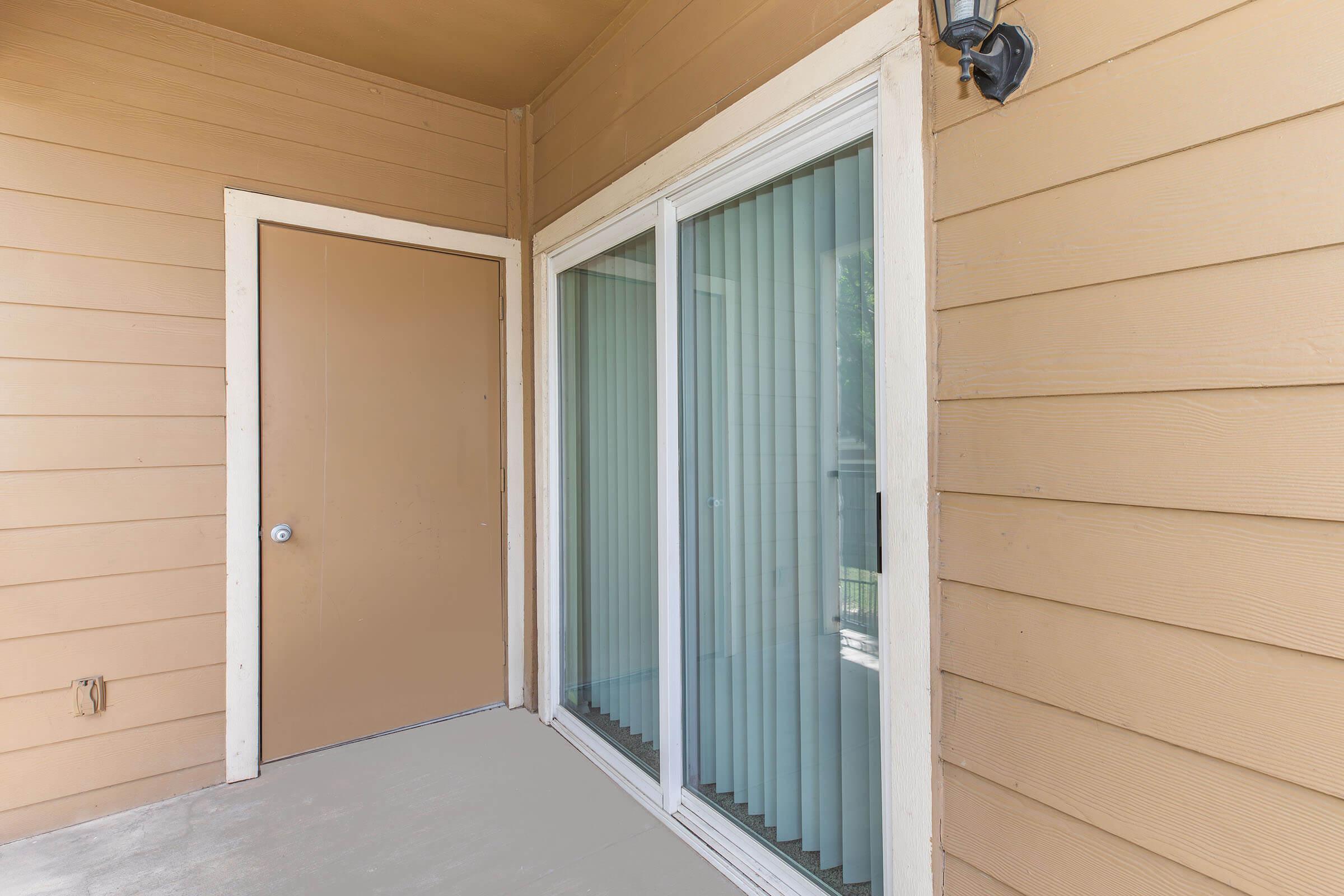 A close-up view of a porch featuring a brown door and large sliding glass window with vertical blinds. The walls are painted in a warm, light brown color, and the floor appears to be a light concrete. A light fixture is mounted above the door. The space is well-lit, indicating a welcoming entrance.