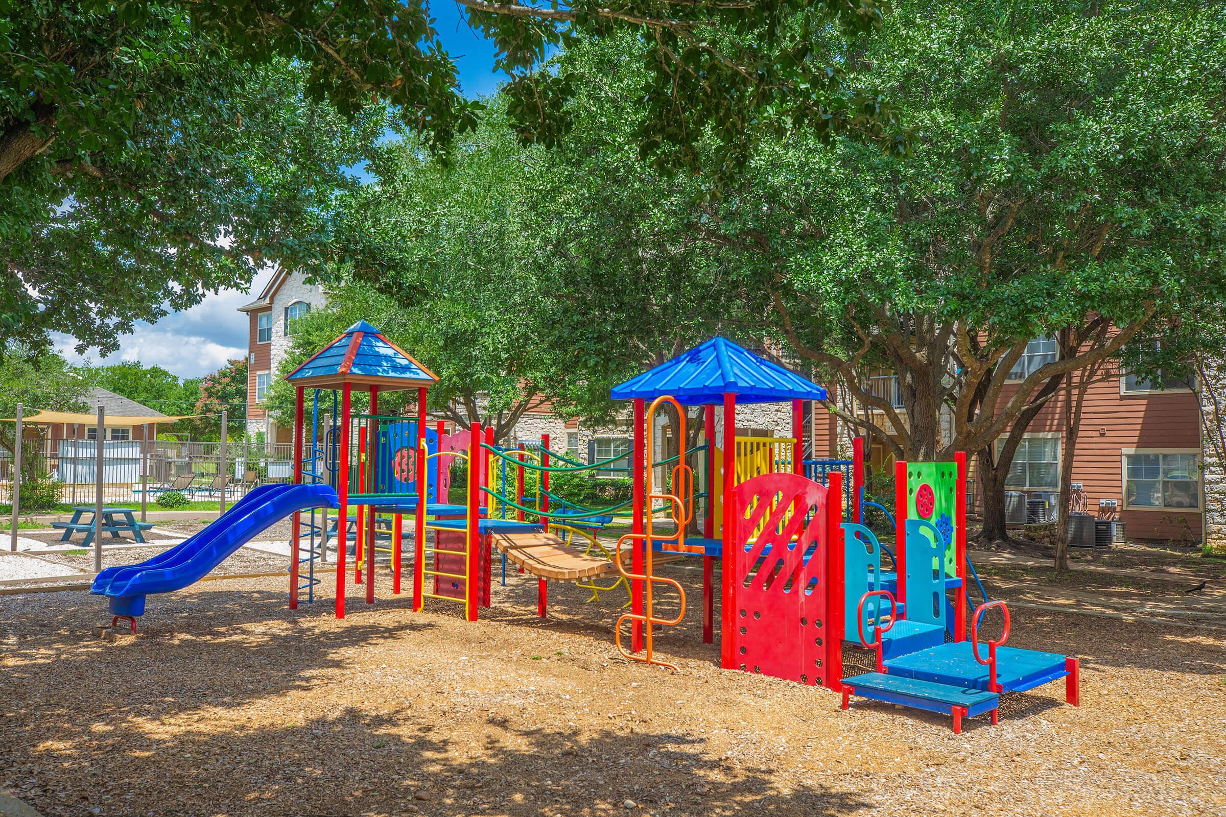 A colorful playground featuring slides, climbing structures, and swings, surrounded by lush green trees. The area is designed for children's play, with equipment in vibrant shades of red and blue, set on a bed of wood chips and near residential buildings. Bright and inviting outdoor space for recreation.