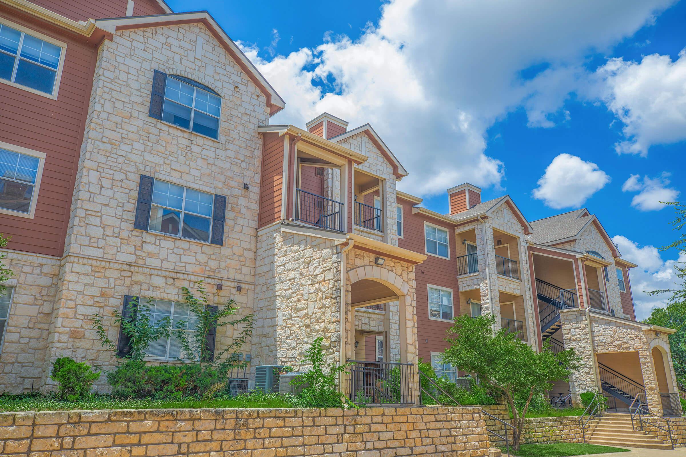 Exterior view of a multi-family apartment building with stone and wooden siding. The structure features multiple balconies, windows, and greenery in front. A blue sky with fluffy clouds complements the scene. The design suggests a modern residential community.
