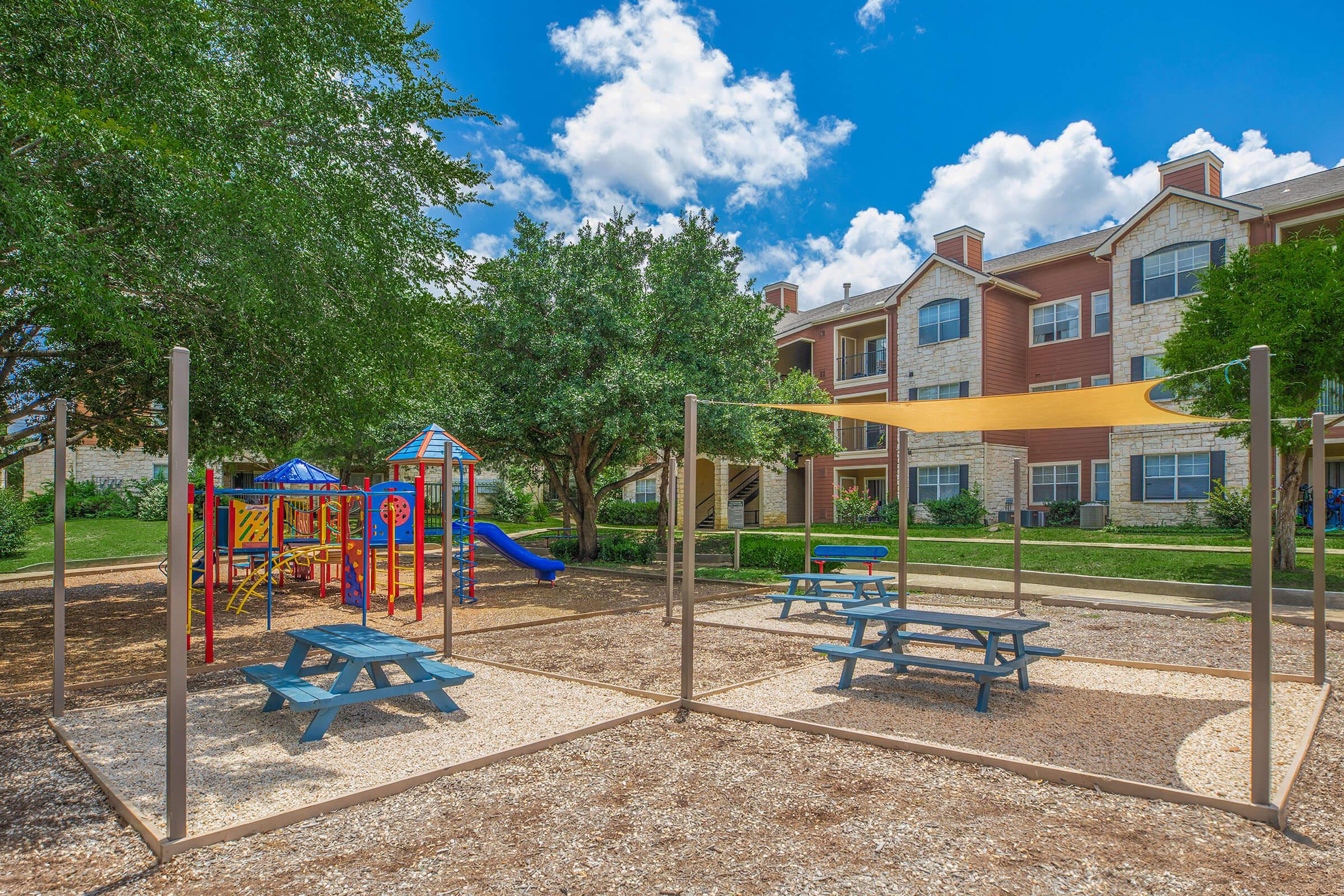 A colorful playground with slides and climbing structures is situated in a grassy area surrounded by trees. There are several picnic tables, and in the background, a multi-story apartment building is visible under a blue sky with fluffy white clouds.