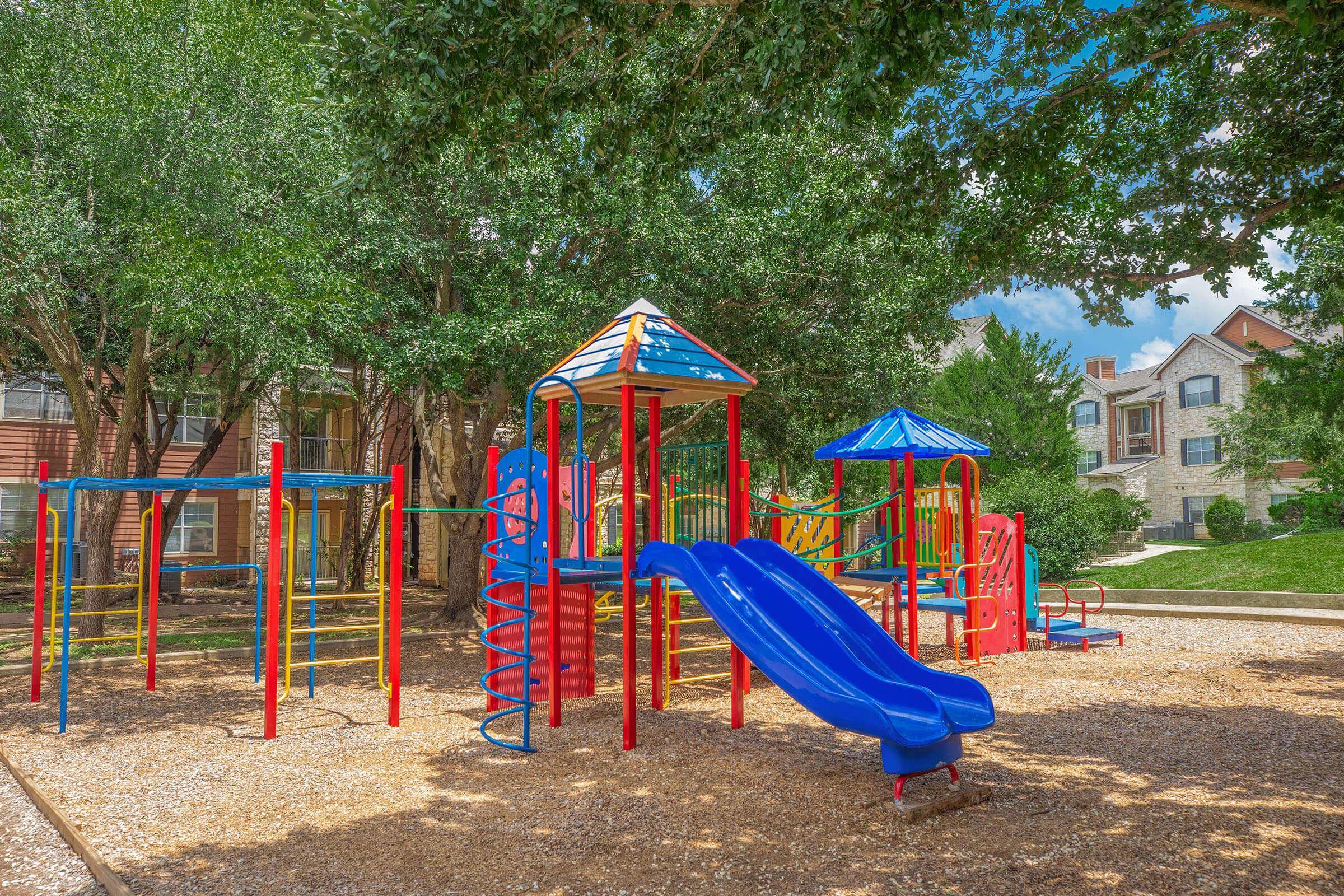 Colorful playground equipment featuring slides, climbing structures, and swings, located in a shaded area with trees, surrounded by a gravel surface. In the background, there are residential buildings and a grassy hillside under a blue sky with scattered clouds.