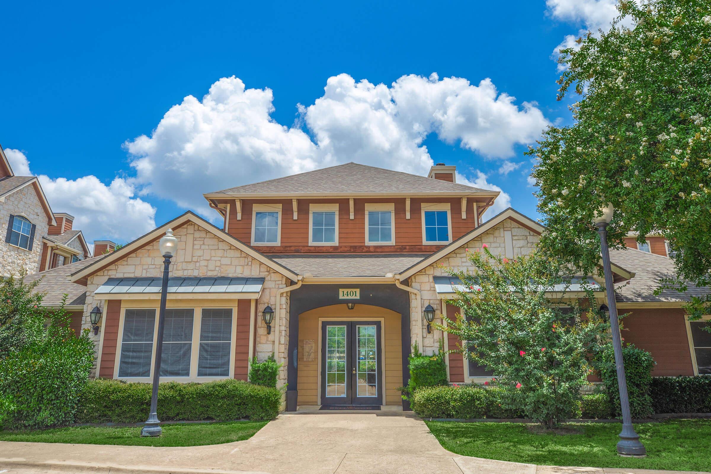 A well-maintained residential building with a charming facade, featuring a combination of stone and wood elements. The front entrance is framed by large windows and surrounded by landscaped greenery, under a blue sky with fluffy clouds. Street lamps are positioned along the walkway.