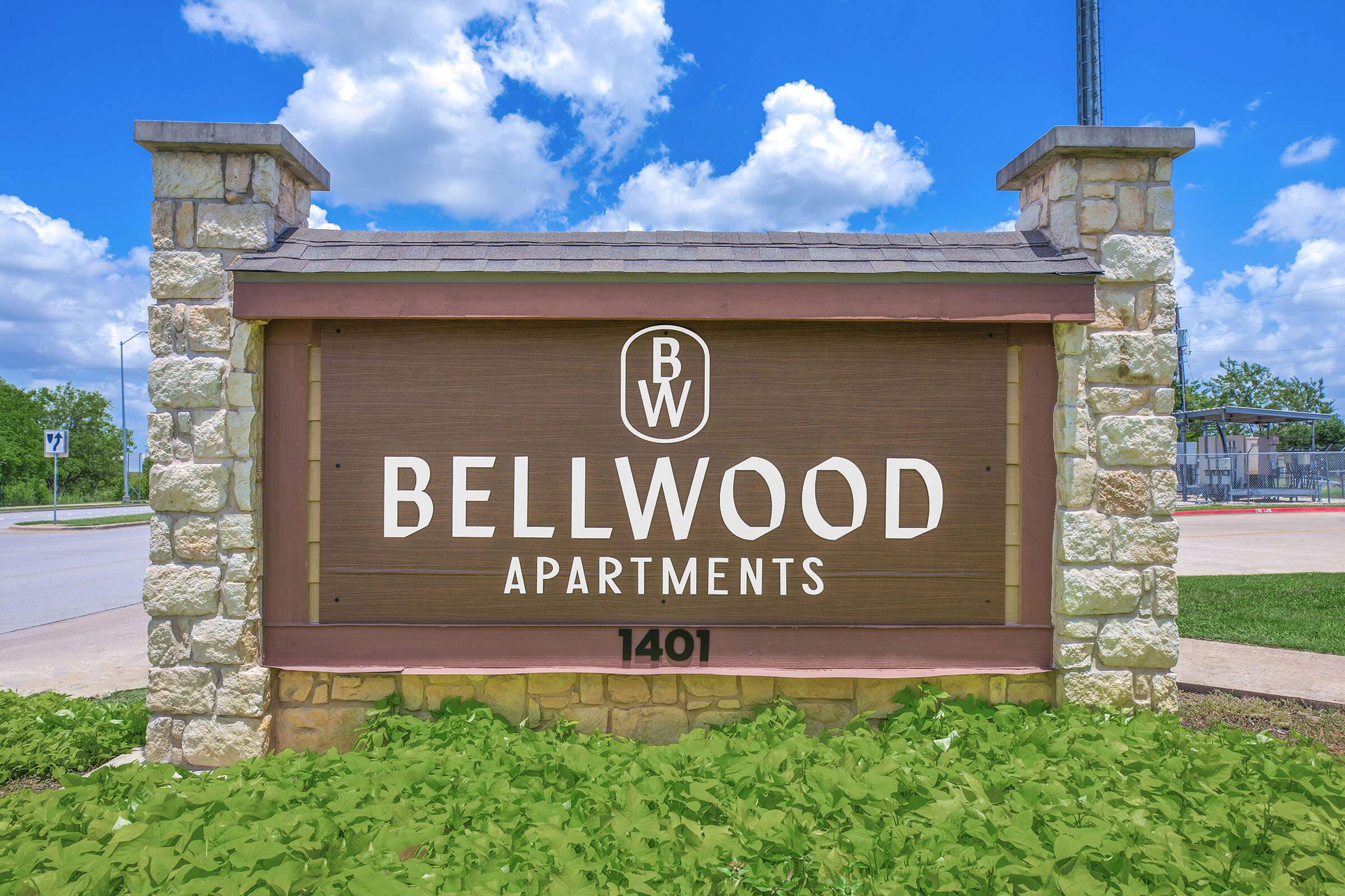 A sign for Bellwood Apartments featuring the name "BELLWOOD APARTMENTS" in large white letters on a wooden background. The sign is bordered with stone, and the location address "1401" is displayed below. The setting includes blue skies with clouds and lush green landscaping.