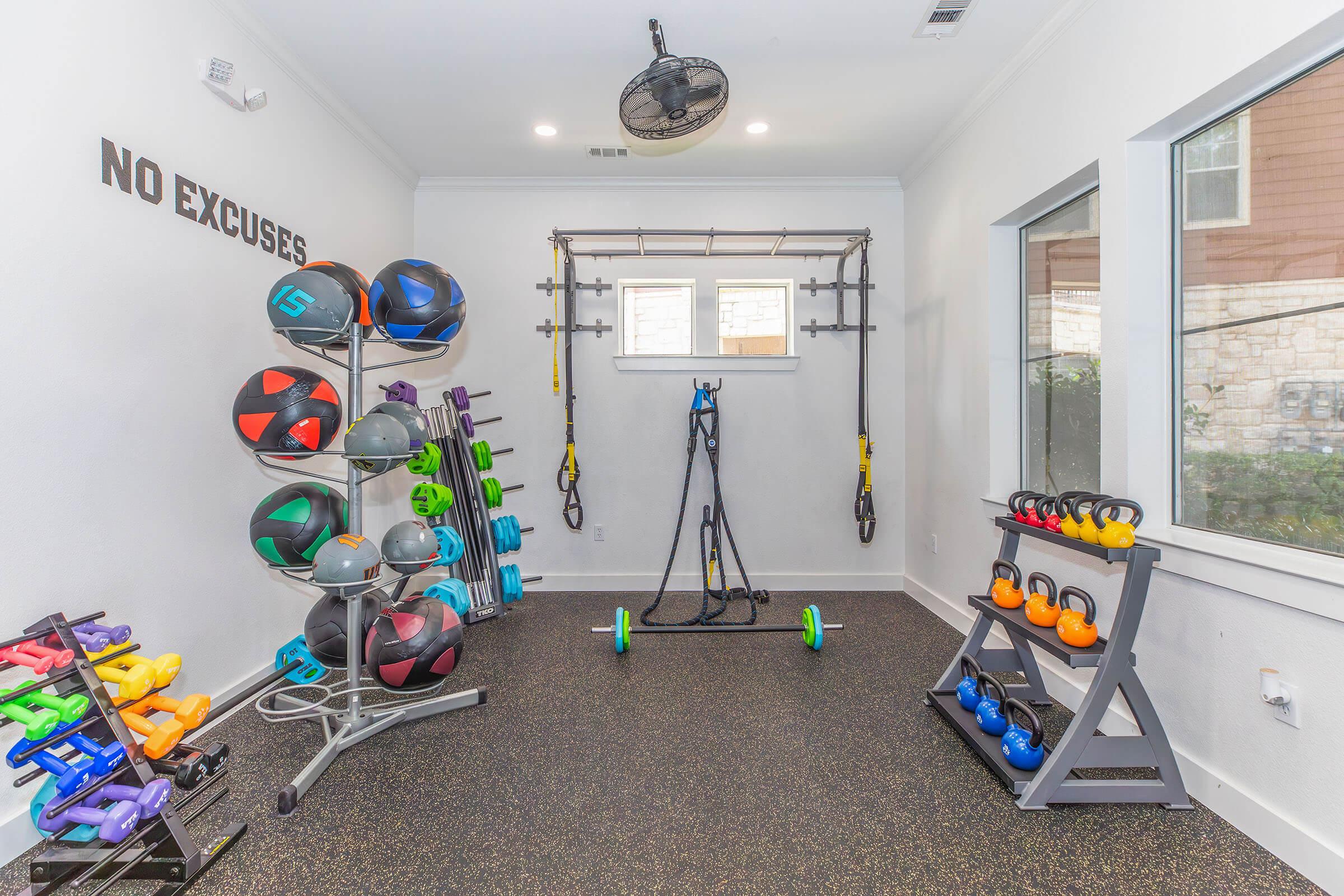 A modern home gym featuring a variety of fitness equipment, including kettlebells, medicine balls, and resistance bands. The walls are decorated with the phrase "NO EXCUSES." Large windows provide natural light, creating an inviting workout space. A ceiling fan adds ventilation.