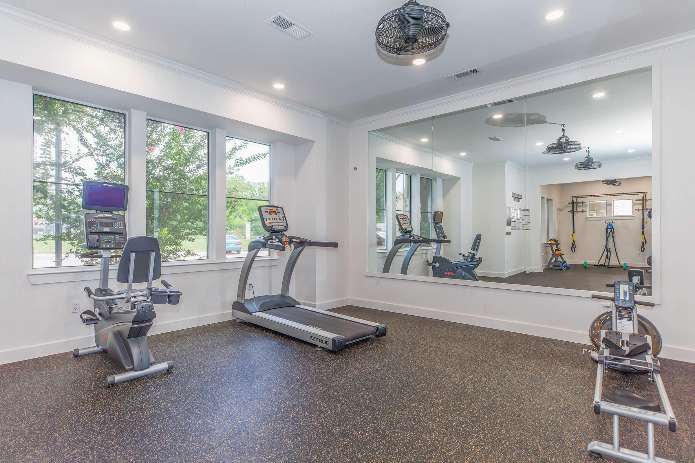 A clean and modern gym space featuring exercise equipment including a treadmill, stationary bike, and weight bench. Large windows provide natural light, and a mirror reflects the equipment. The flooring is dark and textured, creating a functional and appealing workout environment.