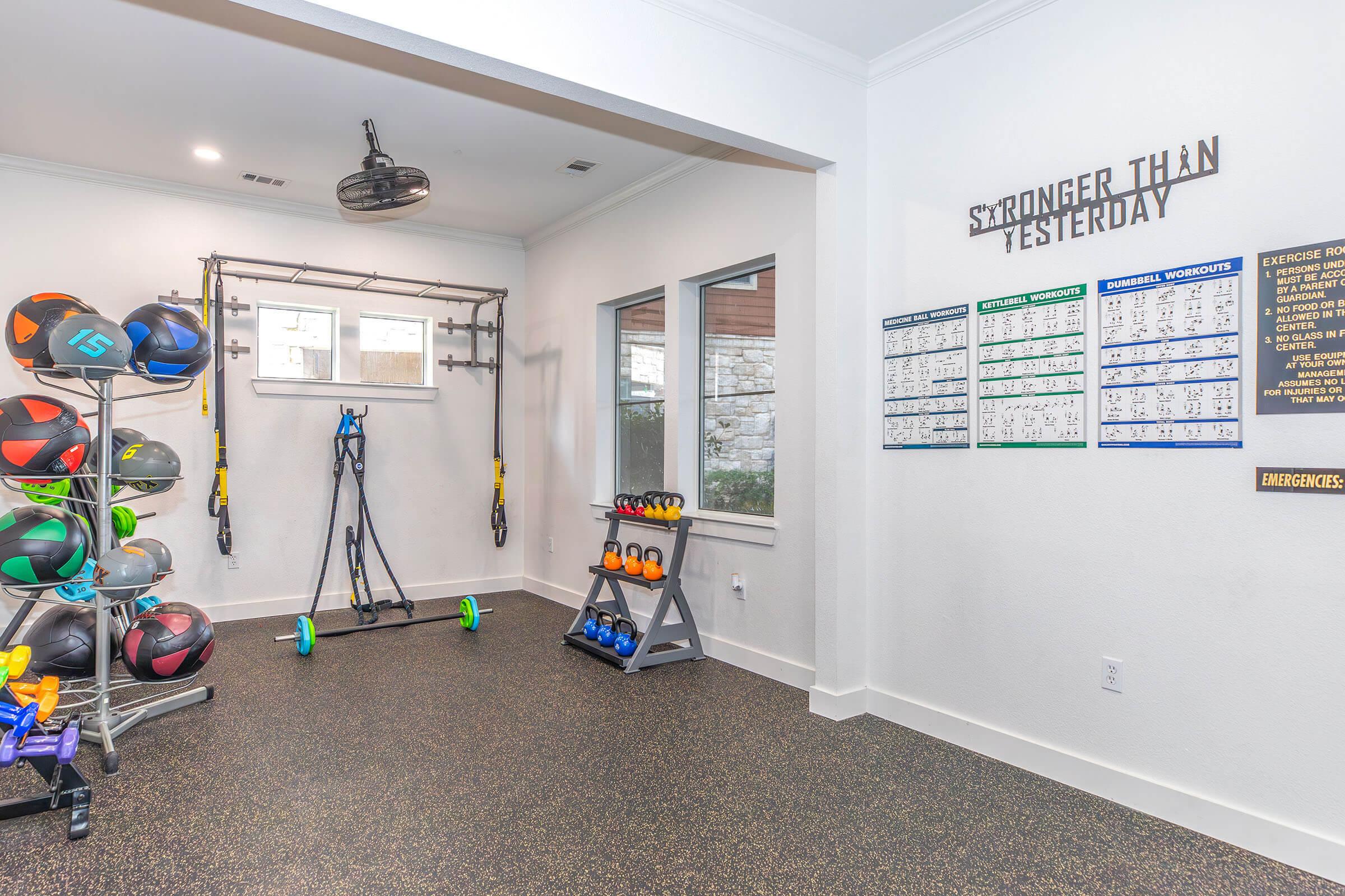 A modern fitness room featuring a variety of exercise equipment, including colorful kettlebells, medicine balls on racks, resistance bands, and a wall-mounted exercise chart. The room has white walls, a black floor, and a motivational sign that says "Stronger Than Yesterday." Natural light comes through two windows.