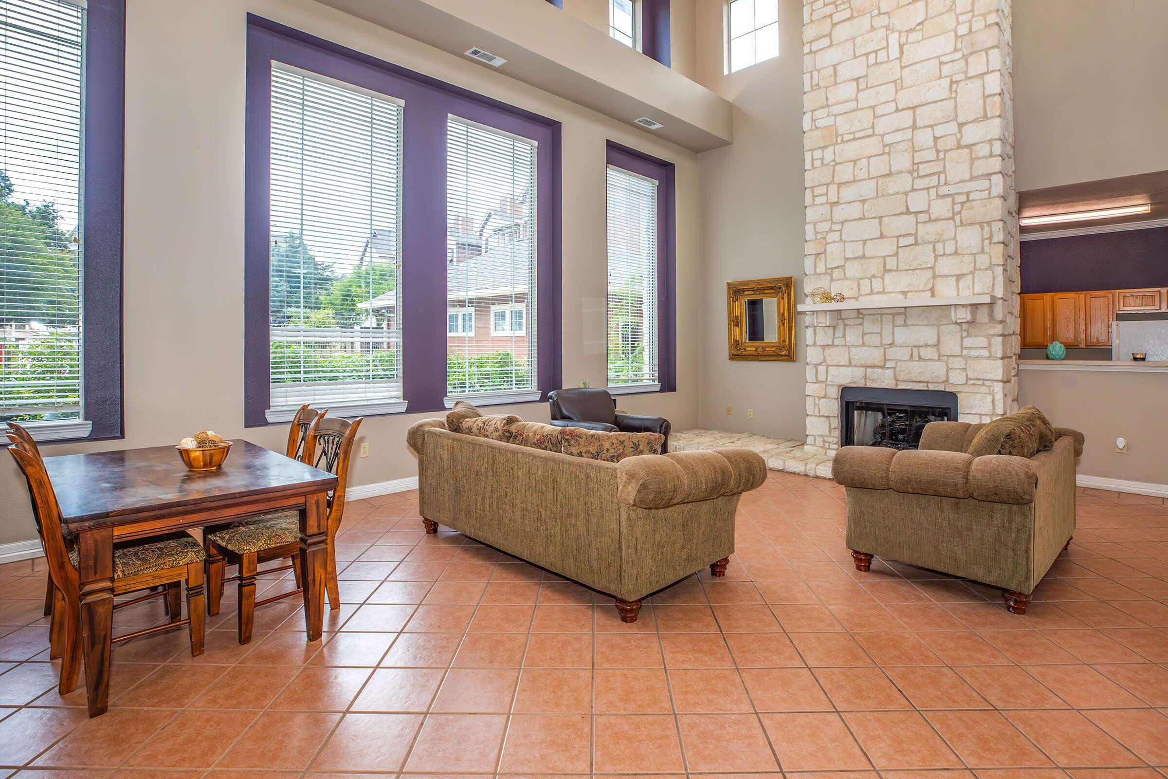 Spacious living room featuring large windows, a stone fireplace, and a cozy seating area with green sofas. A wooden dining table with chairs is positioned nearby, creating a warm and inviting atmosphere. Tile flooring and neutral walls enhance the room's brightness.