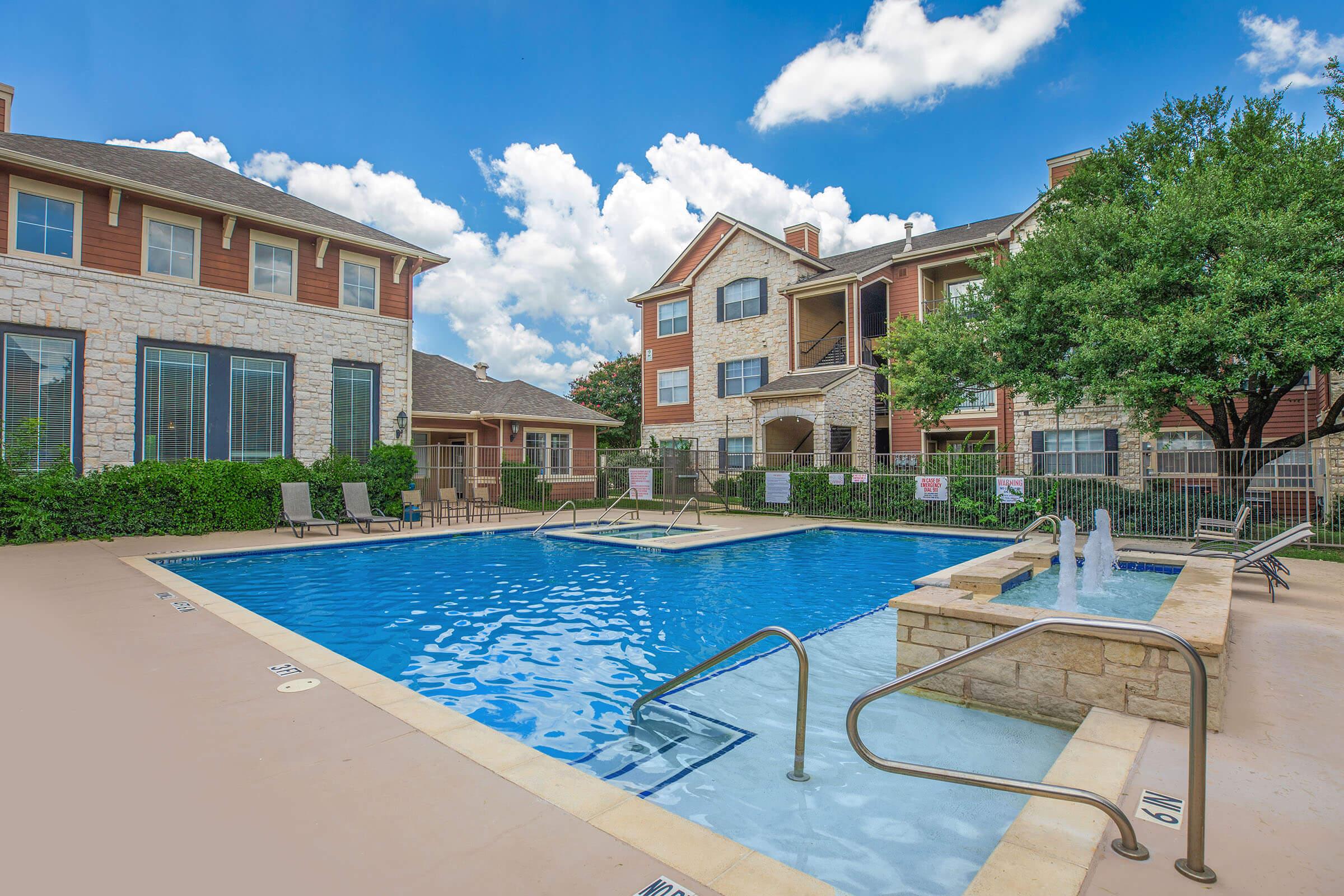 A large swimming pool surrounded by lounge chairs and a fountain, with residential buildings in the background. The sky is bright blue with fluffy white clouds, and well-maintained greenery frames the area. The pool area looks inviting and is designed for relaxation and leisure.