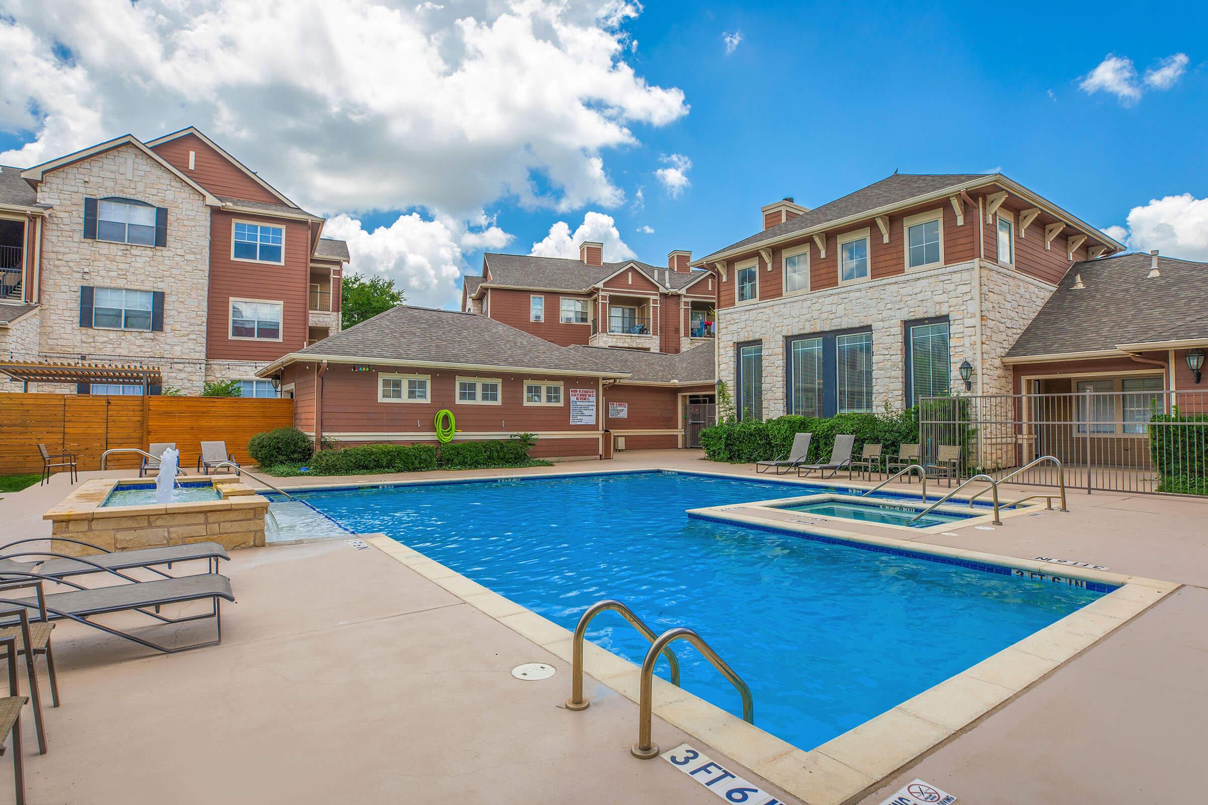 A bright outdoor swimming pool surrounded by lounge chairs, situated in a residential area. The pool area features a stone deck, a fenced perimeter, and is adjacent to a two-story building with multiple windows. The sky is partly cloudy, adding a cheerful atmosphere.