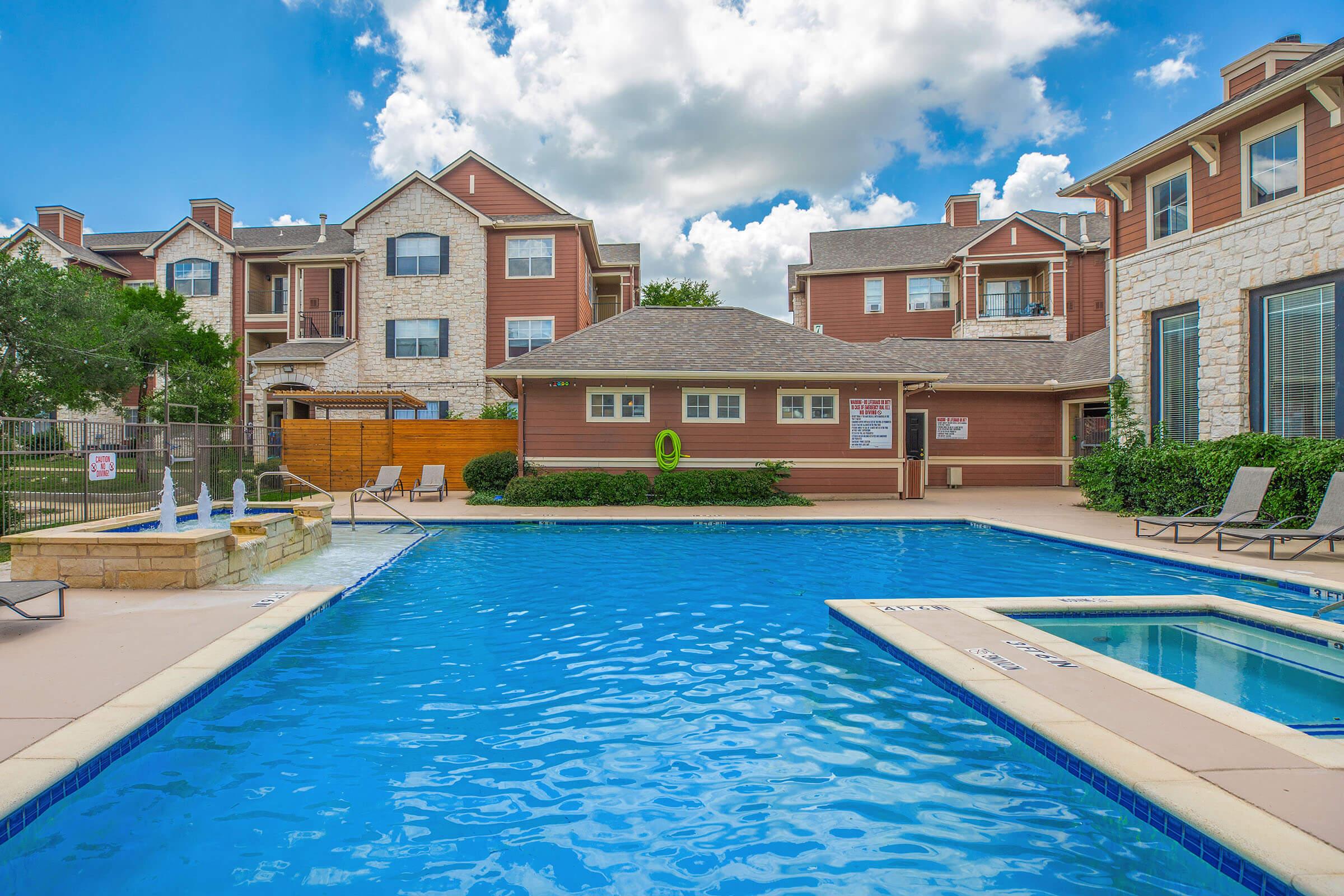 A swimming pool surrounded by lounge chairs, with a hot tub nearby. The area features a light-colored building with multiple stories in the background and a fence. The sky is bright blue with scattered clouds.