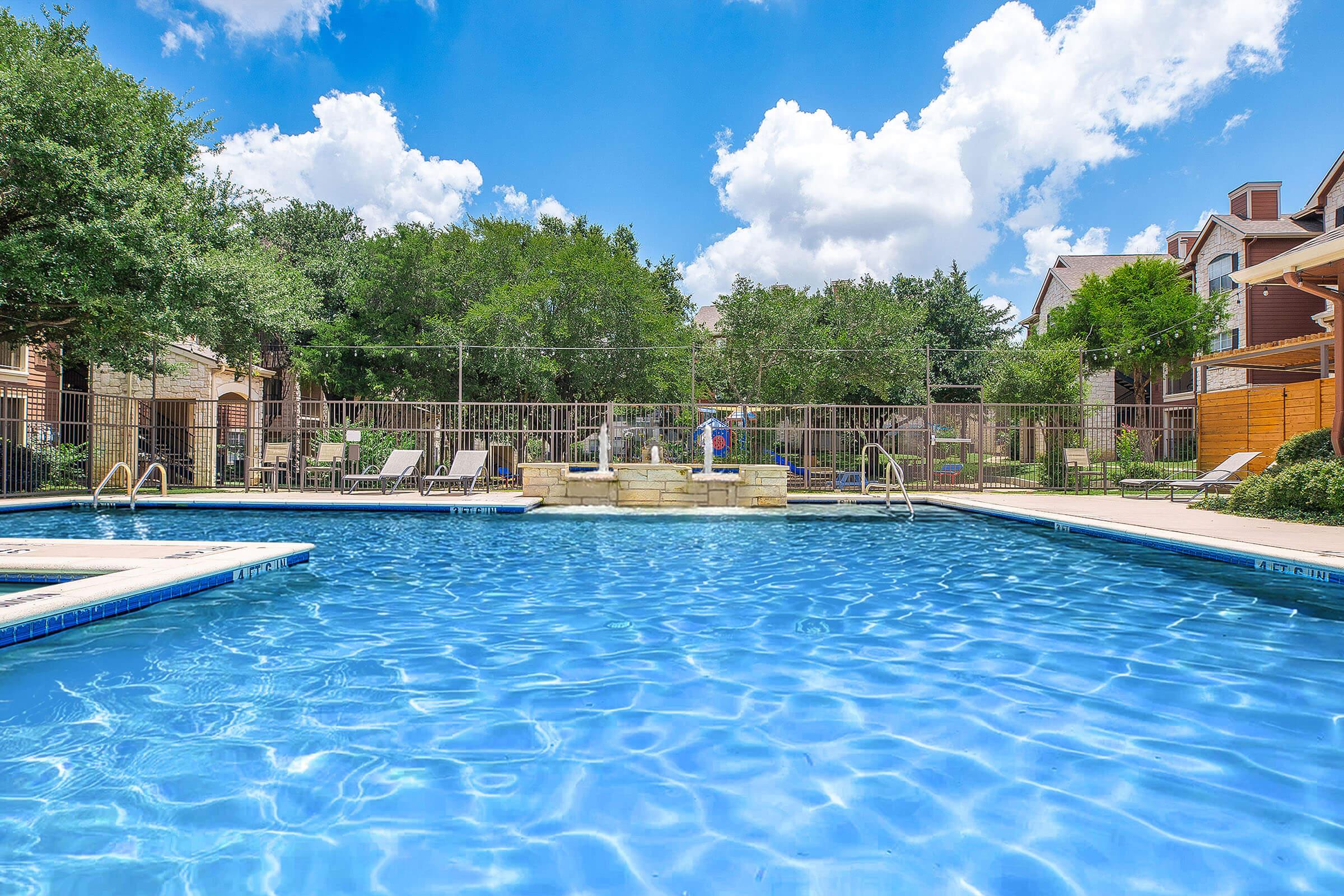 A sunny outdoor swimming pool surrounded by lounge chairs and green trees. Clear blue water reflects the sky with fluffy white clouds. A small stone fountain is visible in the center of the pool area, creating a relaxing atmosphere. A fence encloses the pool for privacy.