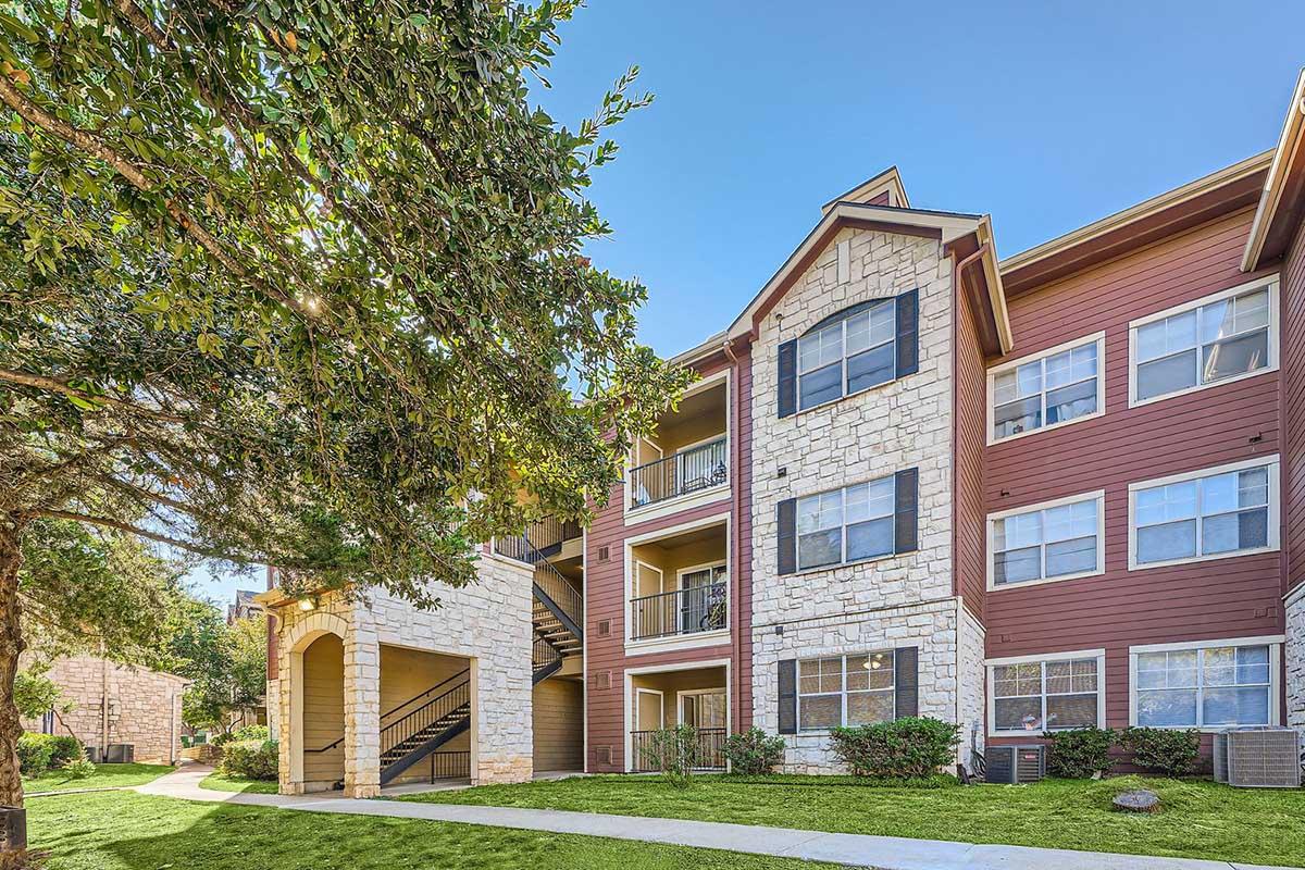 A well-maintained apartment building with a mix of stone and siding exteriors. The building features multiple balconies, large windows, and a landscaped lawn with trees and pathway. Clear blue sky overhead adds to the inviting atmosphere.