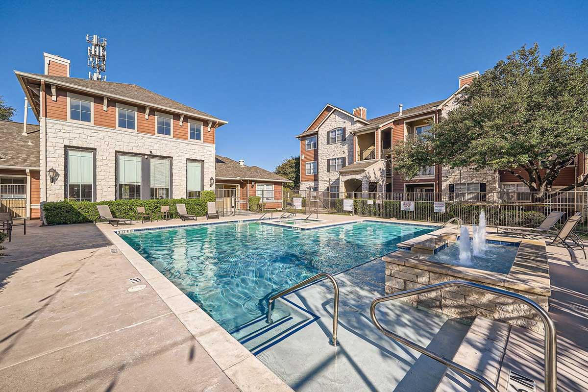 A clear view of a swimming pool surrounded by residential buildings. The pool features a stone fountain and is equipped with steps for entry. Lounge chairs line the pool area, and trees provide shade in the background under a bright blue sky. The scene conveys a relaxing outdoor space.