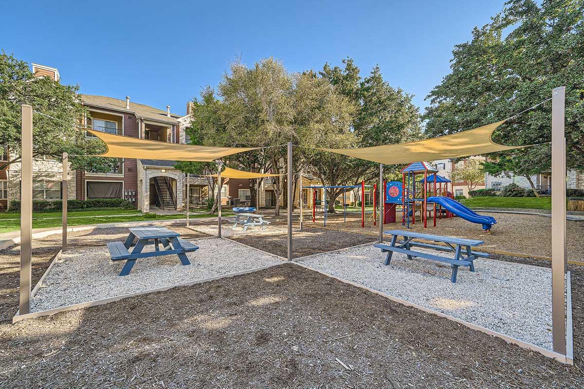 Playground area with blue picnic tables and playground equipment, including a slide and climbing structure, shaded by large canopies. Surrounding grass and trees provide a green space, with apartment buildings visible in the background under a clear blue sky.