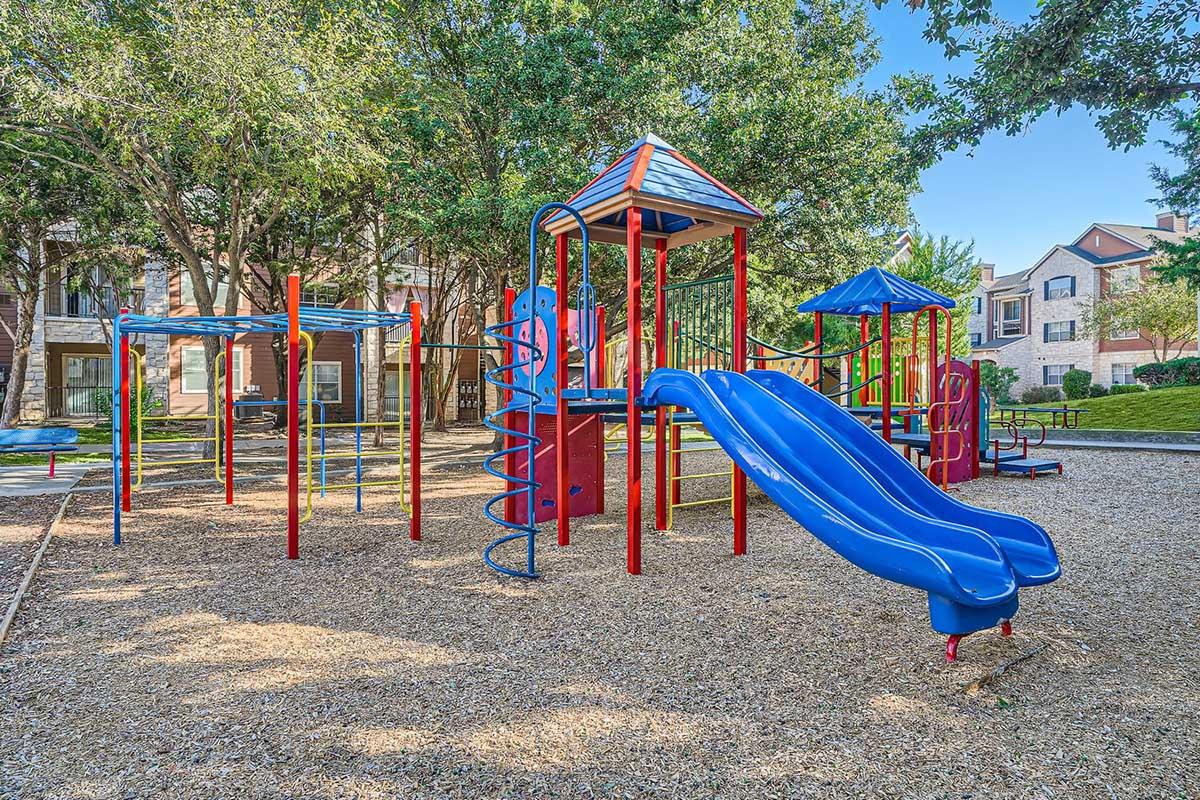 A vibrant playground with colorful equipment, including blue slides, climbing structures, and swings, set on a gravel surface surrounded by trees and residential buildings in the background. The scene is sunny and inviting, perfect for children to play and explore.