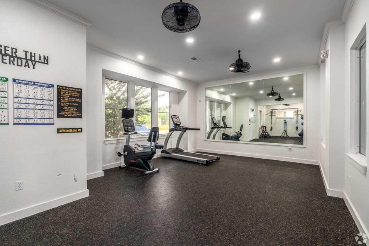 A bright and modern gym space featuring exercise equipment including a treadmill and exercise bike. The room has large windows for natural light, mirrors on one wall, and motivational signage. The flooring is dark, and there are ceiling fans for ventilation.