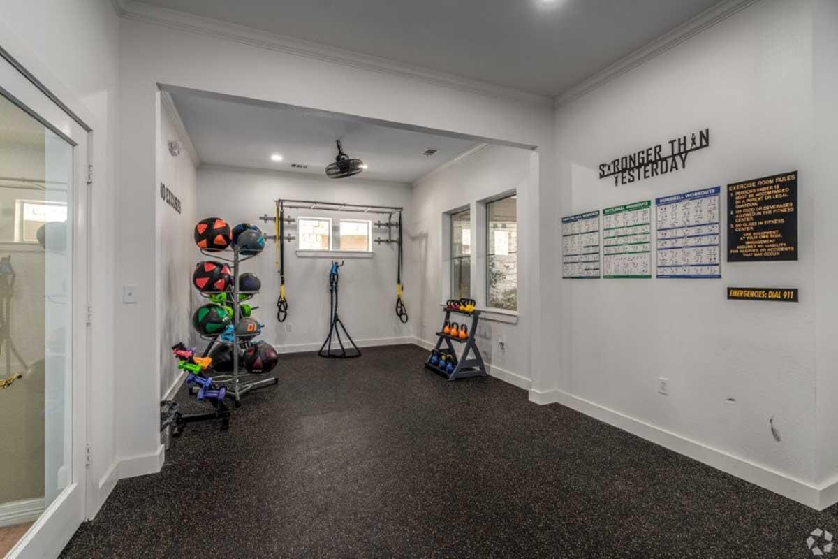 A modern home gym featuring various fitness equipment, including medicine balls, resistance bands, and weight training accessories. There's a motivational wall chart with the phrase "Stronger than yesterday" and large windows allowing natural light into the space.