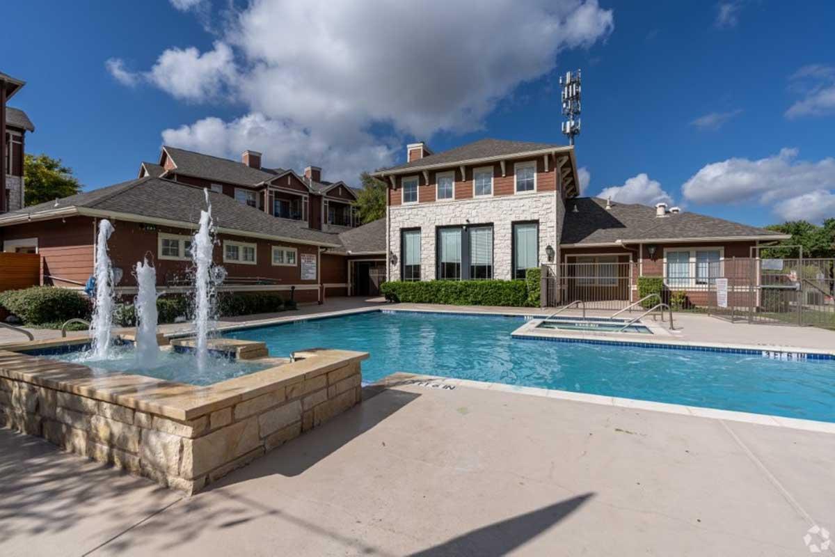 A swimming pool area featuring a clear blue pool, a decorative fountain, and nearby lounge chairs. In the background, there are residential buildings with a stone and brick façade, surrounded by greenery and a blue sky with scattered clouds.