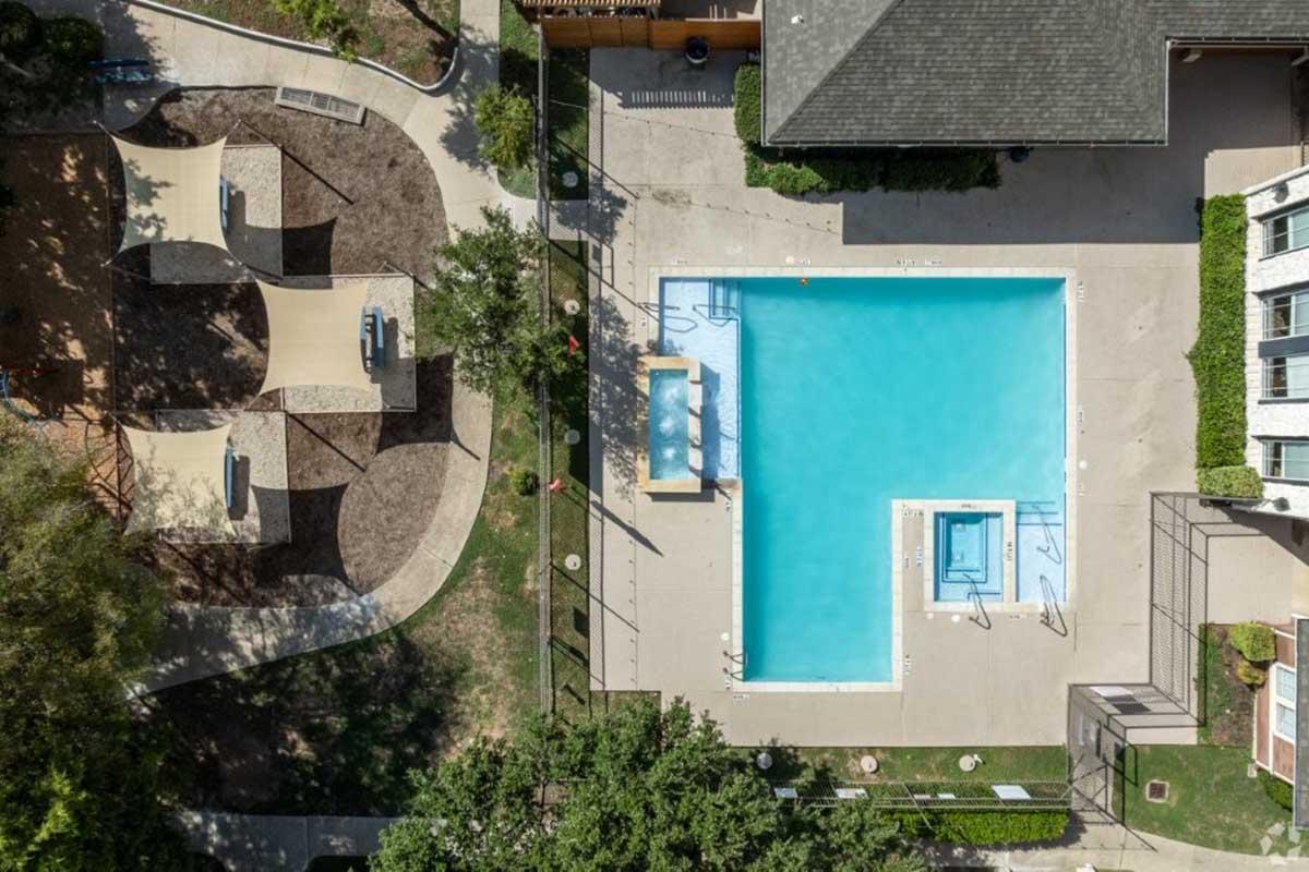 An aerial view of a rectangular swimming pool surrounded by a deck and lounge chairs. Nearby, there are landscaped areas with trees and a playground structure. Walking paths lead around the pool area, featuring benches and shaded spots. The scene is bright and inviting, with clear blue water in the pool.