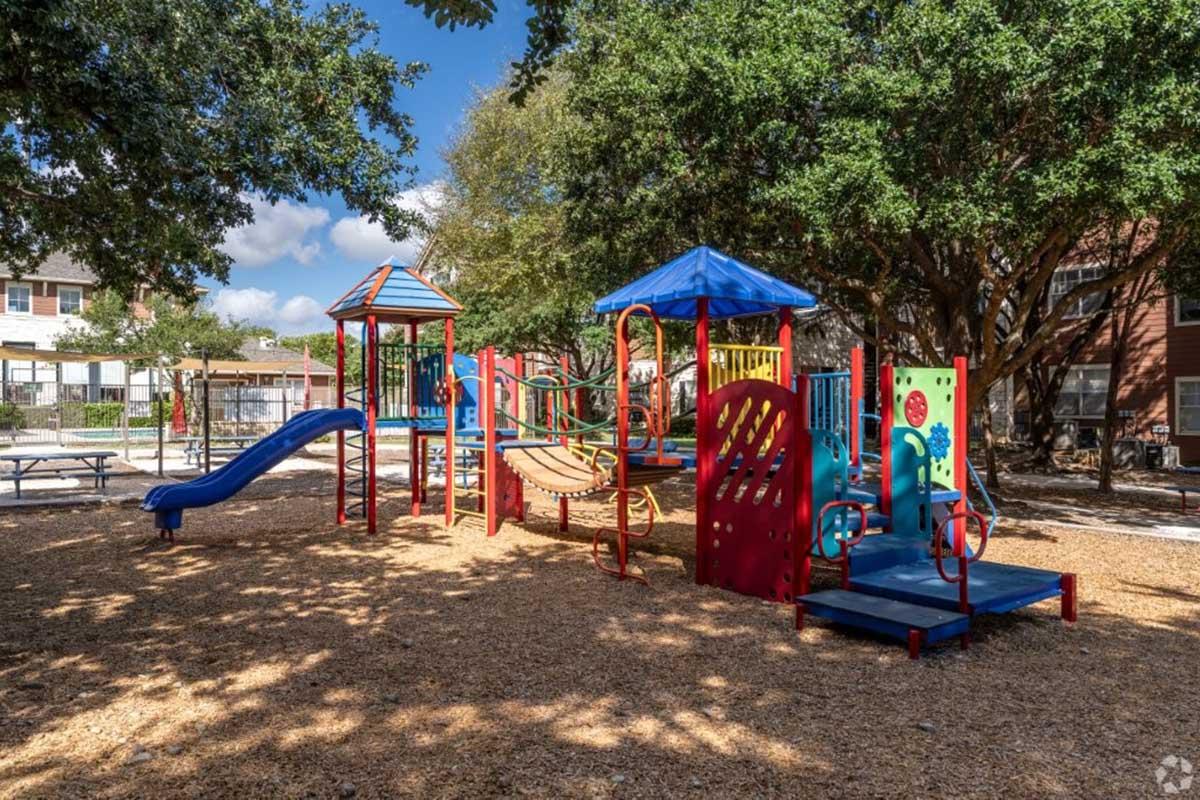 Colorful playground with slides, climbing structures, and benches, surrounded by trees. The ground is covered in wood chips, and the sky is partly cloudy.
