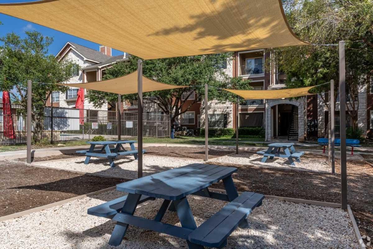 A spacious outdoor area featuring several blue picnic tables shaded by large, yellow canopies. The ground is covered with gravel, and surrounding greenery includes trees and bushes. In the background, there are apartment buildings and areas for relaxation.