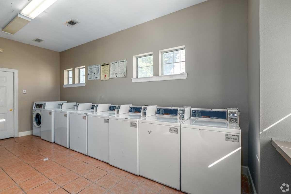 A laundry room featuring multiple white washing machines lined up against a wall. The space has beige walls, several small windows allowing natural light, and a door on the left. There are also information posters on the wall above the machines. The floor is tiled in a light color.